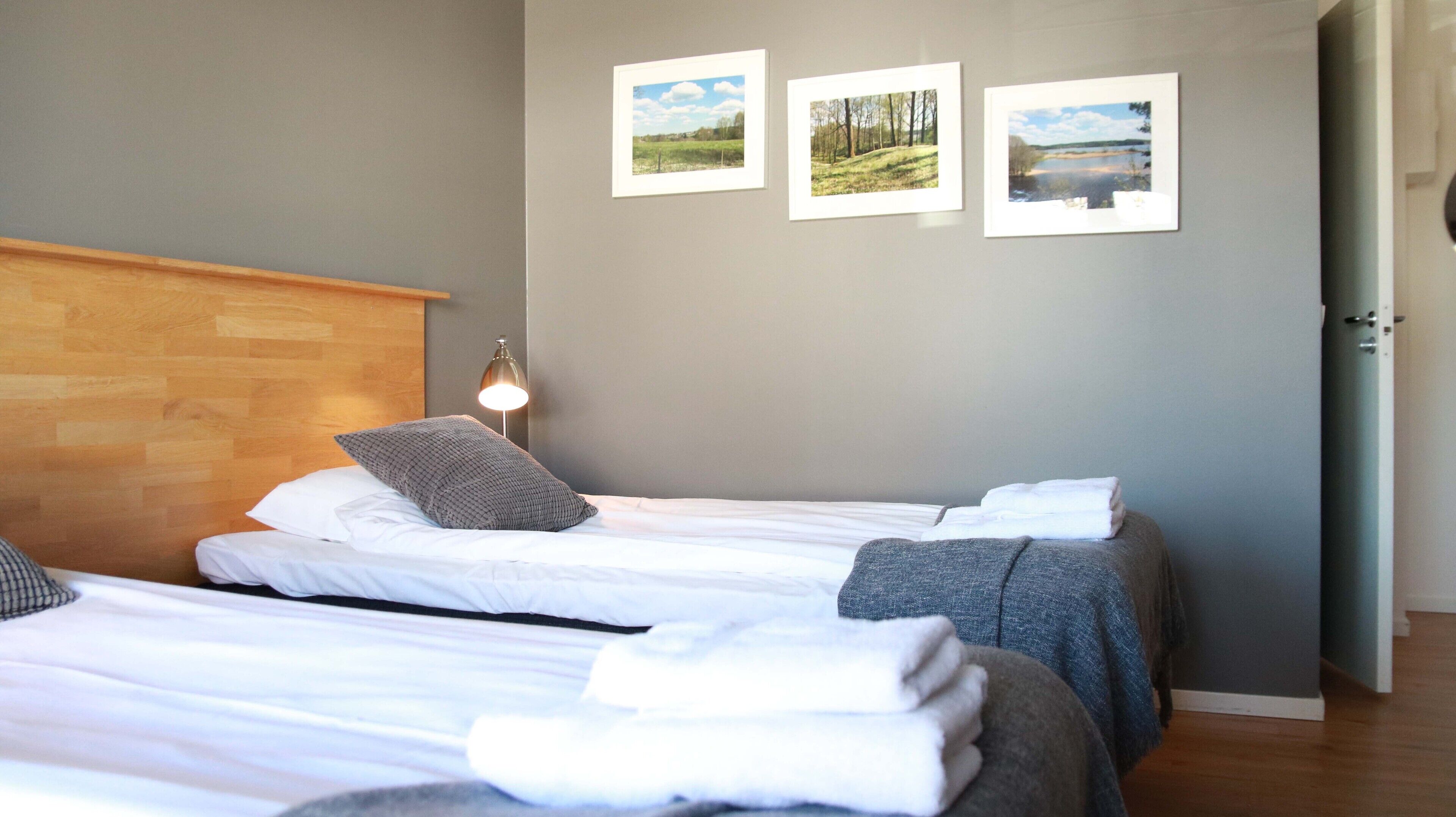 Hotellihuone, jossa on kaksi sänkyä, puinen pääty ja yöpöytälamppu sekä kolme kehystettyä maisemakuvaa seinällä.