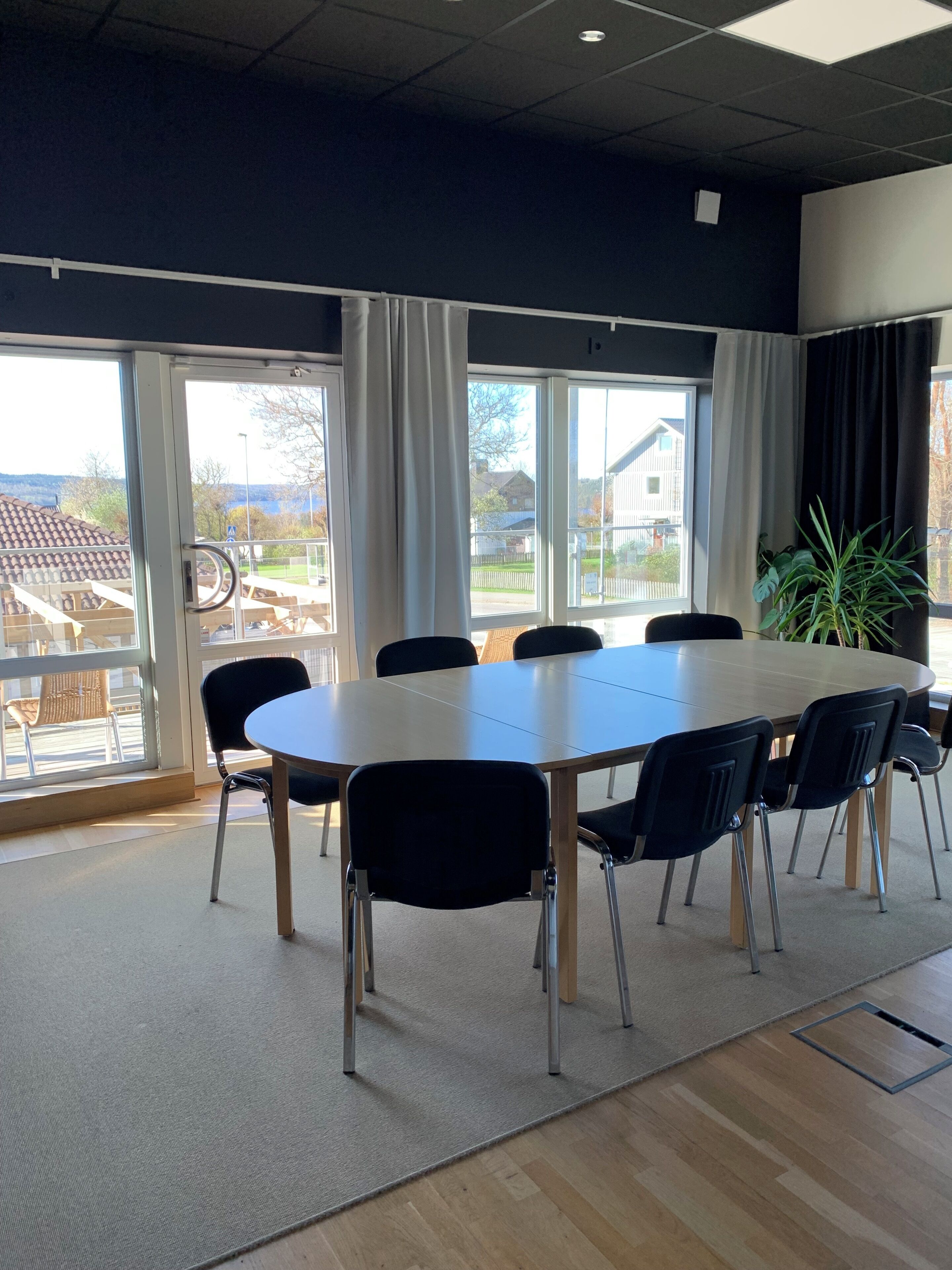 A conference room with a large table and chairs, large windows with curtains, and a view of houses outside.
