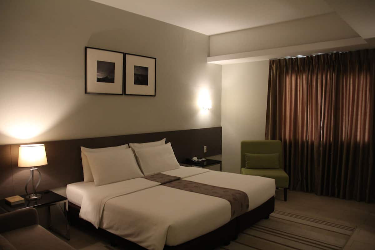 A hotel room with a large bed, a nightstand with a lamp, a green armchair, and two framed pictures on the wall.