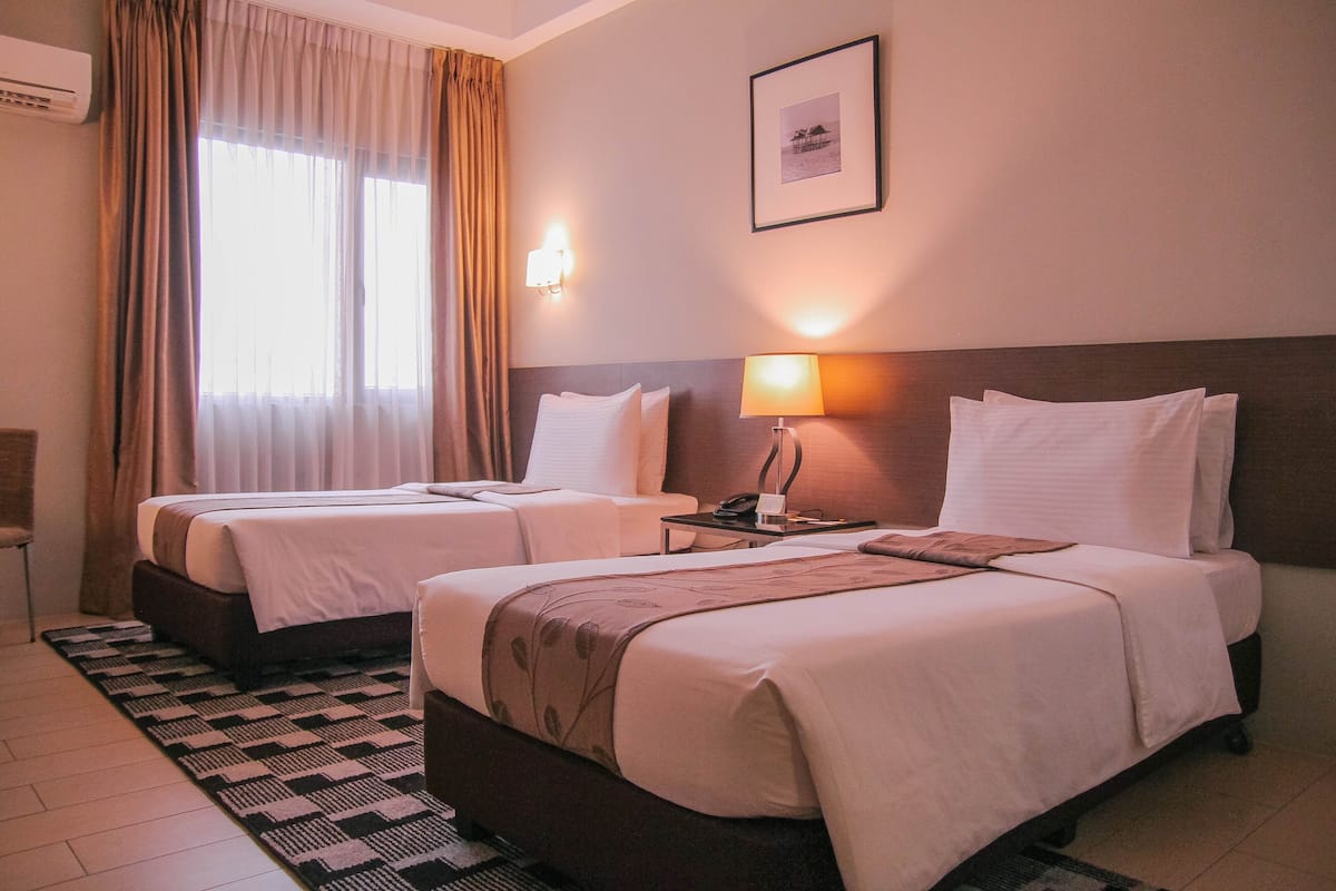 Two single beds with white linens, a bedside table with a lamp, a framed picture on the wall, and a window with curtains.