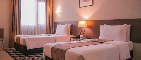 Duas camas de solteiro com roupa de cama branca, uma mesa de cabeceira com abajur, um quadro na parede e uma janela com cortinas.