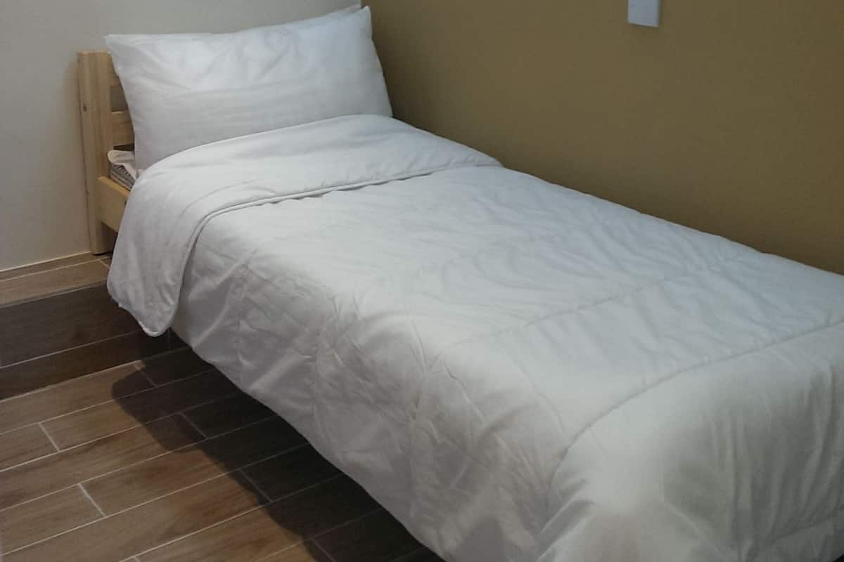 Quarto pequeno com uma cama de solteiro, cabeceira de madeira, parede bege e interruptor de luz.