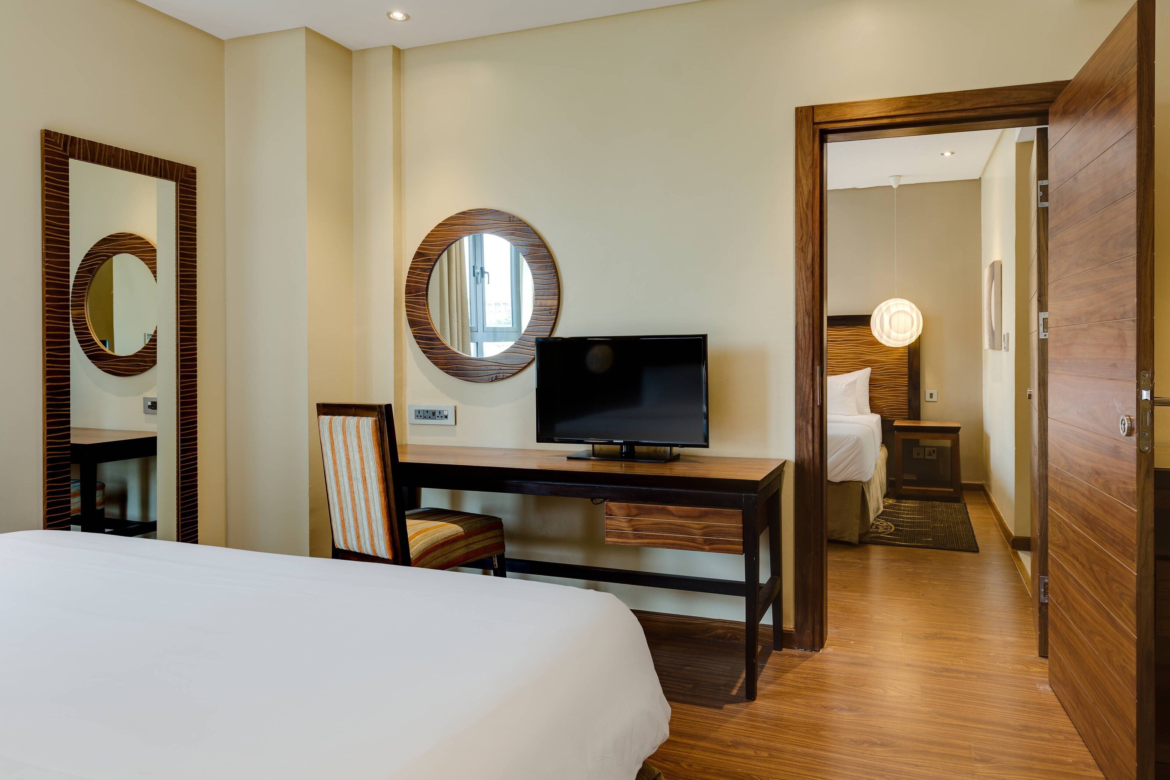 Habitación de hotel con cama, escritorio con televisor, espejo y puerta que da a otra habitación.