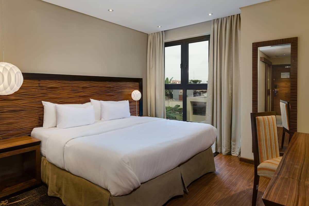 A hotel room with a large bed, a wooden headboard, a desk with a chair, and a large window with curtains.