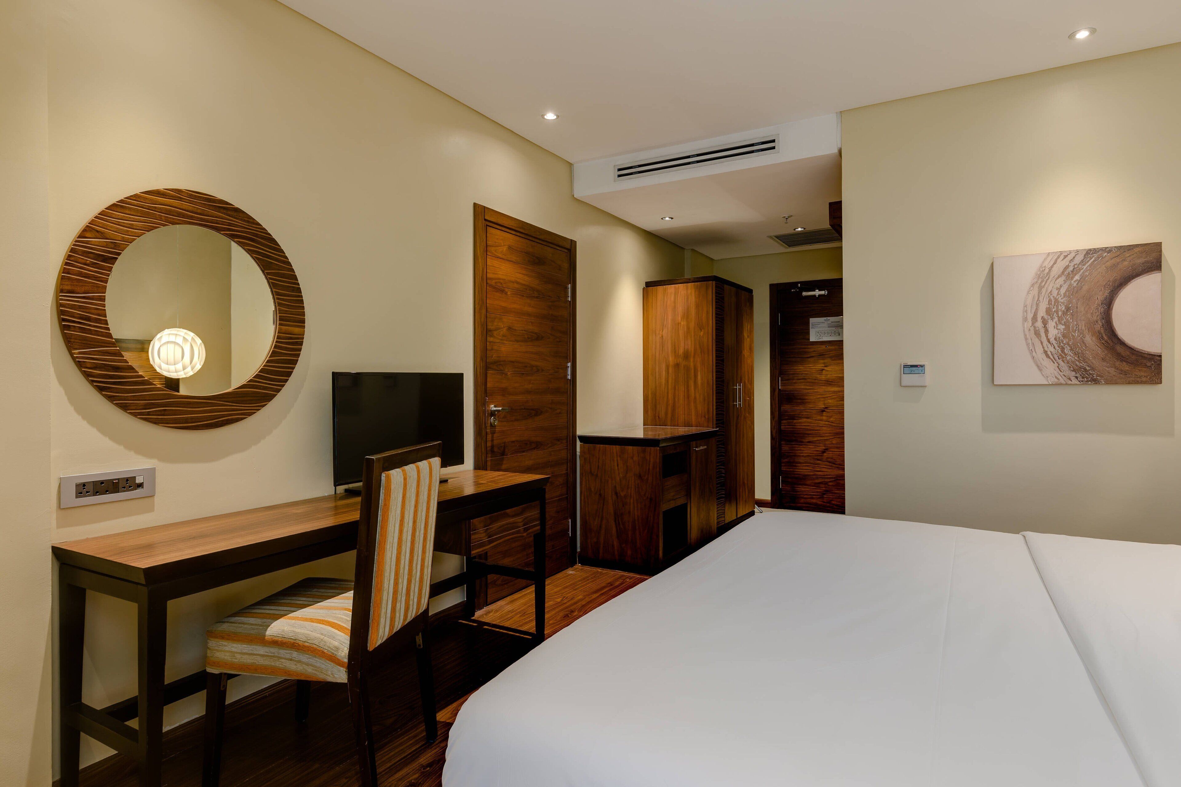 A hotel room with a bed, a desk with a chair, a television, a wardrobe, and a mirror.