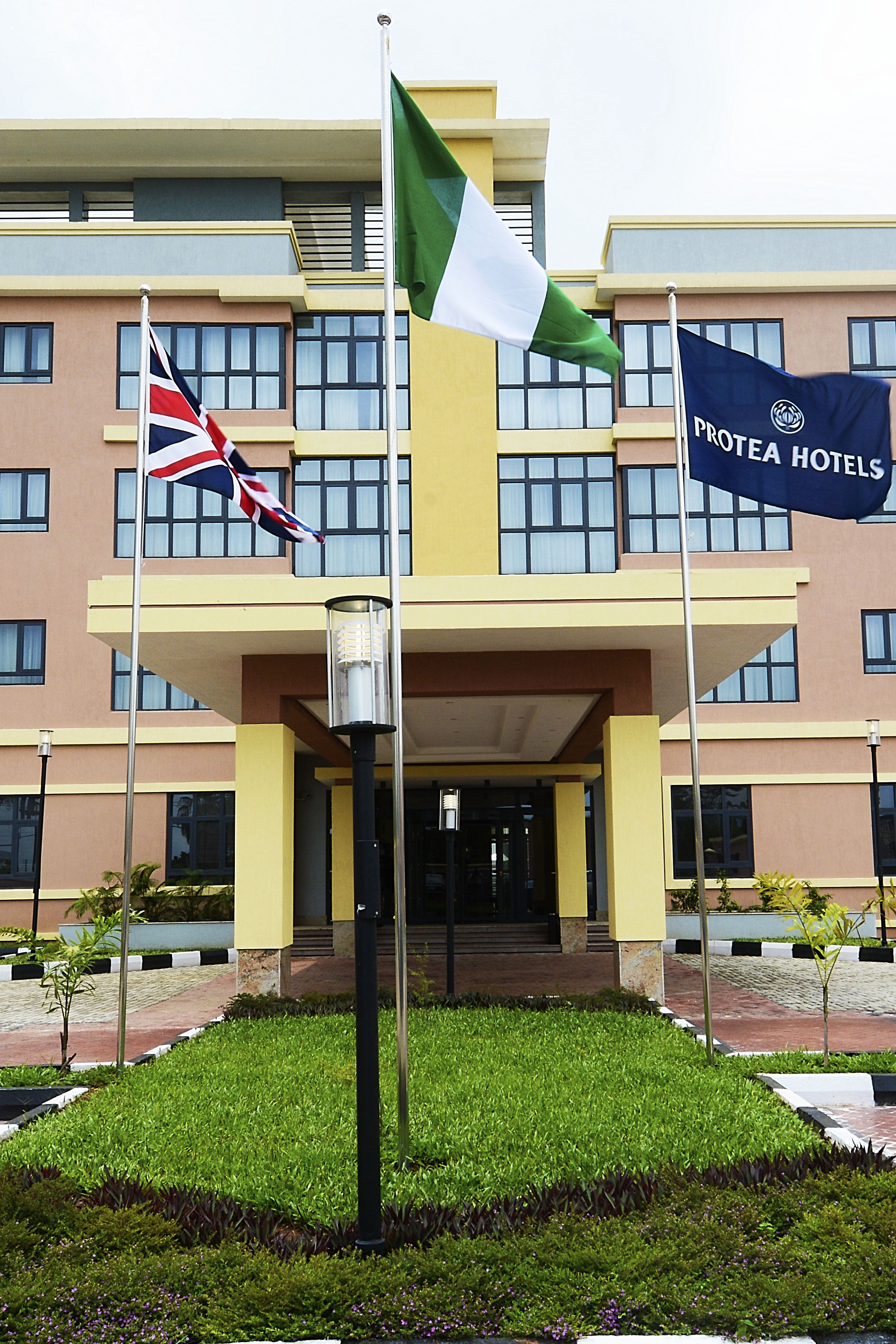 A hotel entrance with a flagpole displaying the Nigerian flag and the Protea Hotels flag.