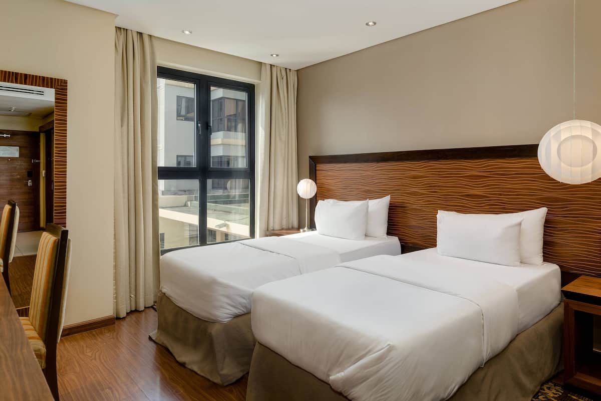 A hotel room with two beds, a wooden headboard, a large window with curtains, a desk with a chair, and a hanging light fixture.
