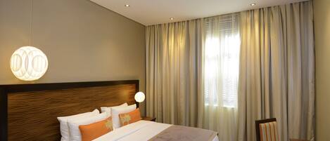 A hotel room with a large bed, a wooden headboard, a bedside table with a lamp, a chair, and a view of the outside through curtains.