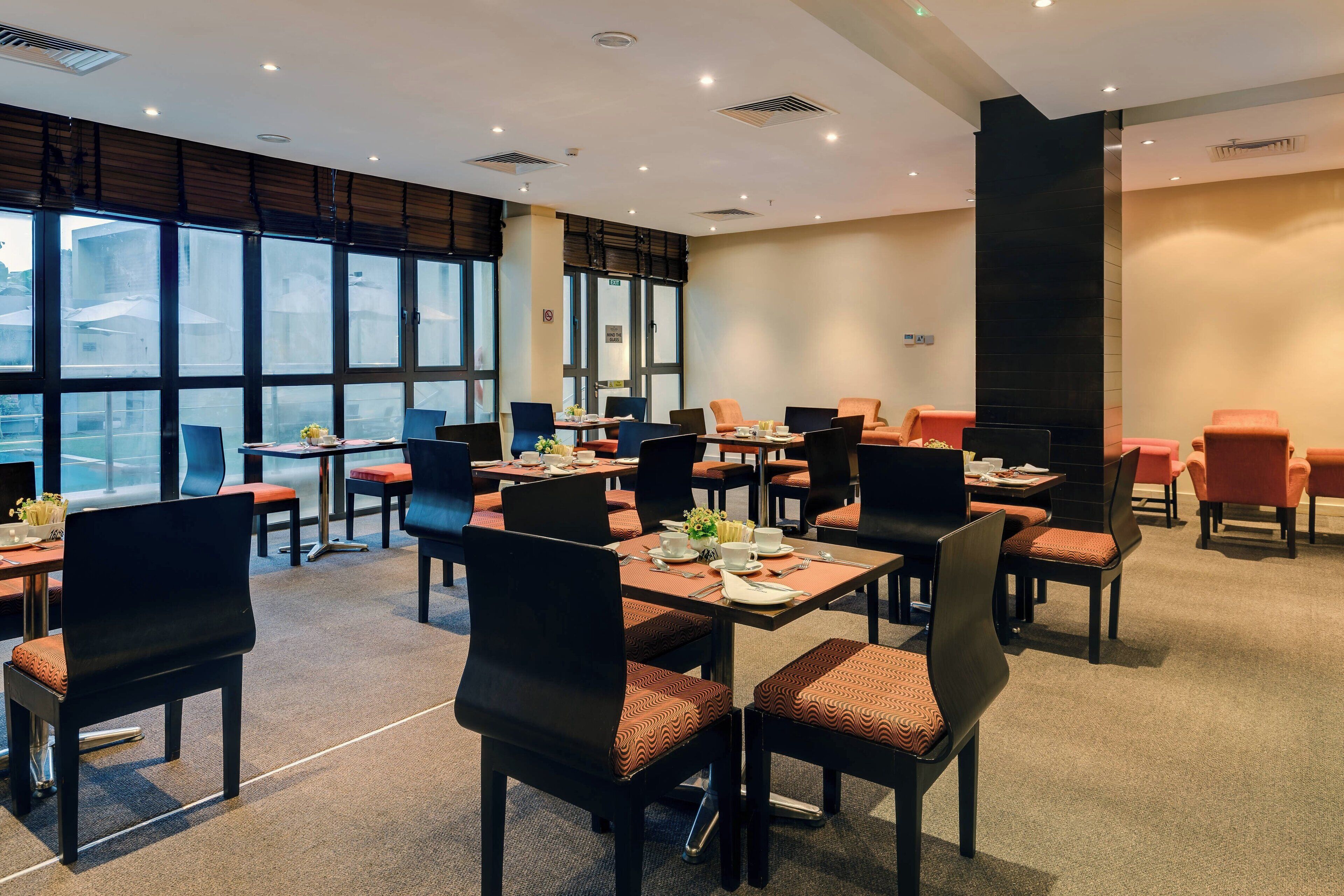 A hotel dining area with multiple tables and chairs, large windows, and a modern interior design.