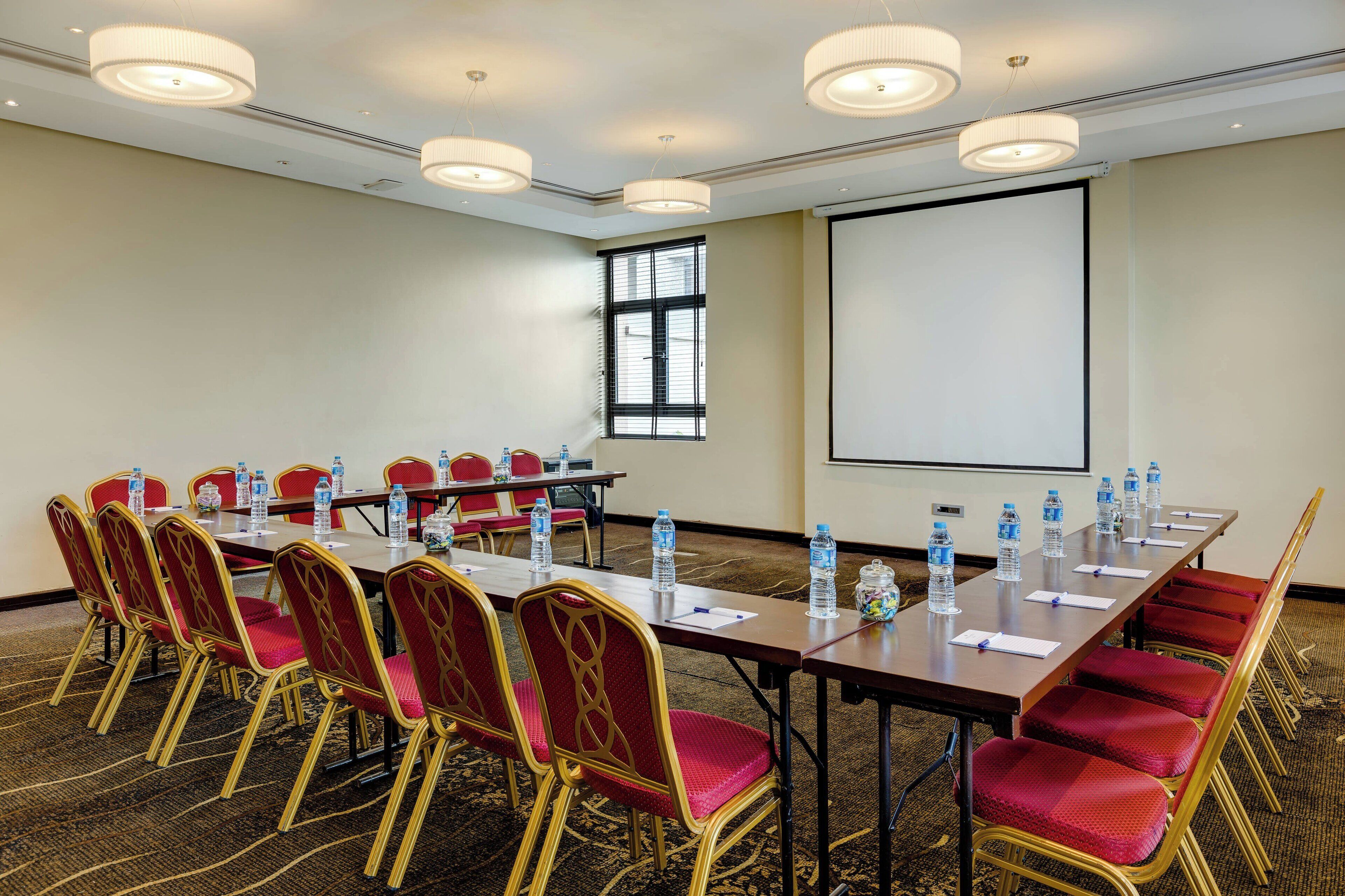 Uma sala de conferências com uma mesa comprida, cadeiras vermelhas, garrafas de água e uma tela de projetor.