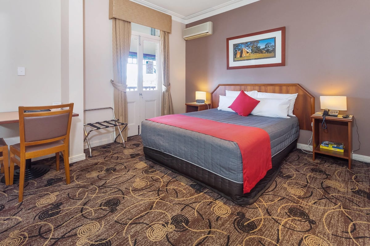 A hotel room with a bed, a chair, a desk, a window with curtains, a painting on the wall, and a patterned carpet.