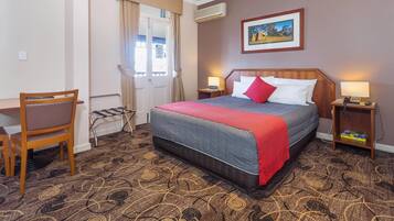 A hotel room with a bed, a chair, a desk, a window with curtains, a painting on the wall, and a patterned carpet.