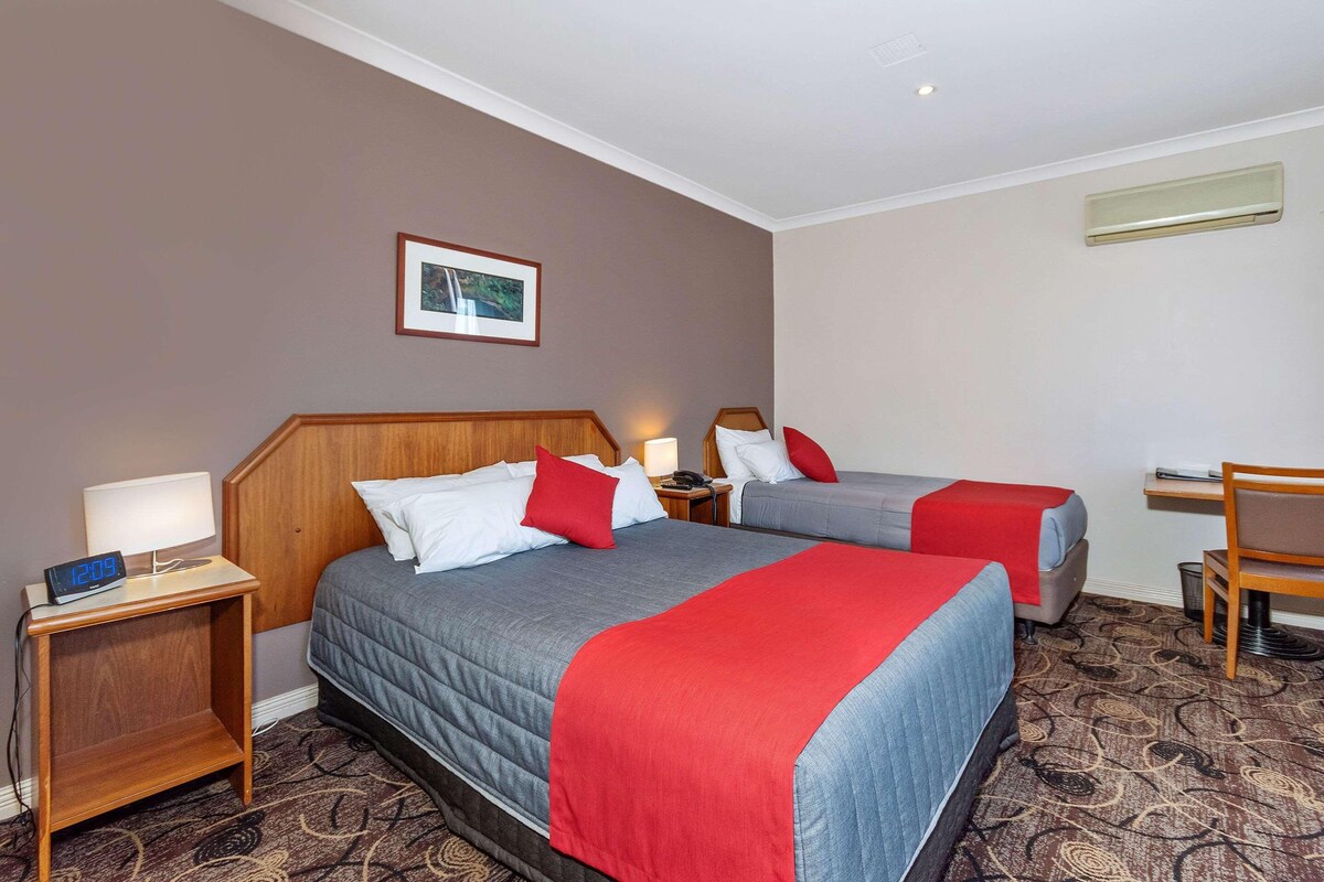 A hotel room with two beds, a desk, a chair, a nightstand with a lamp, and a picture on the wall.