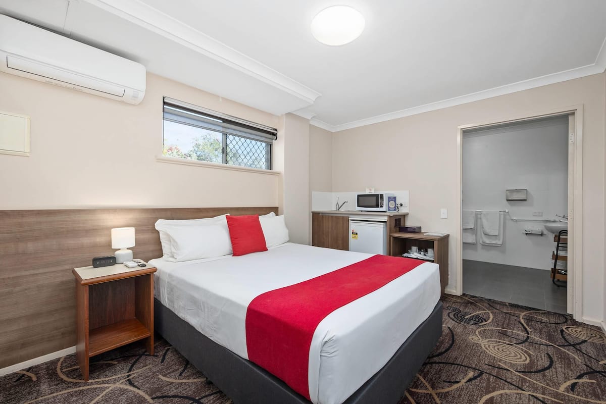 A hotel room with a bed, a nightstand, a desk with a microwave, a small refrigerator, and a bathroom visible through an open door.