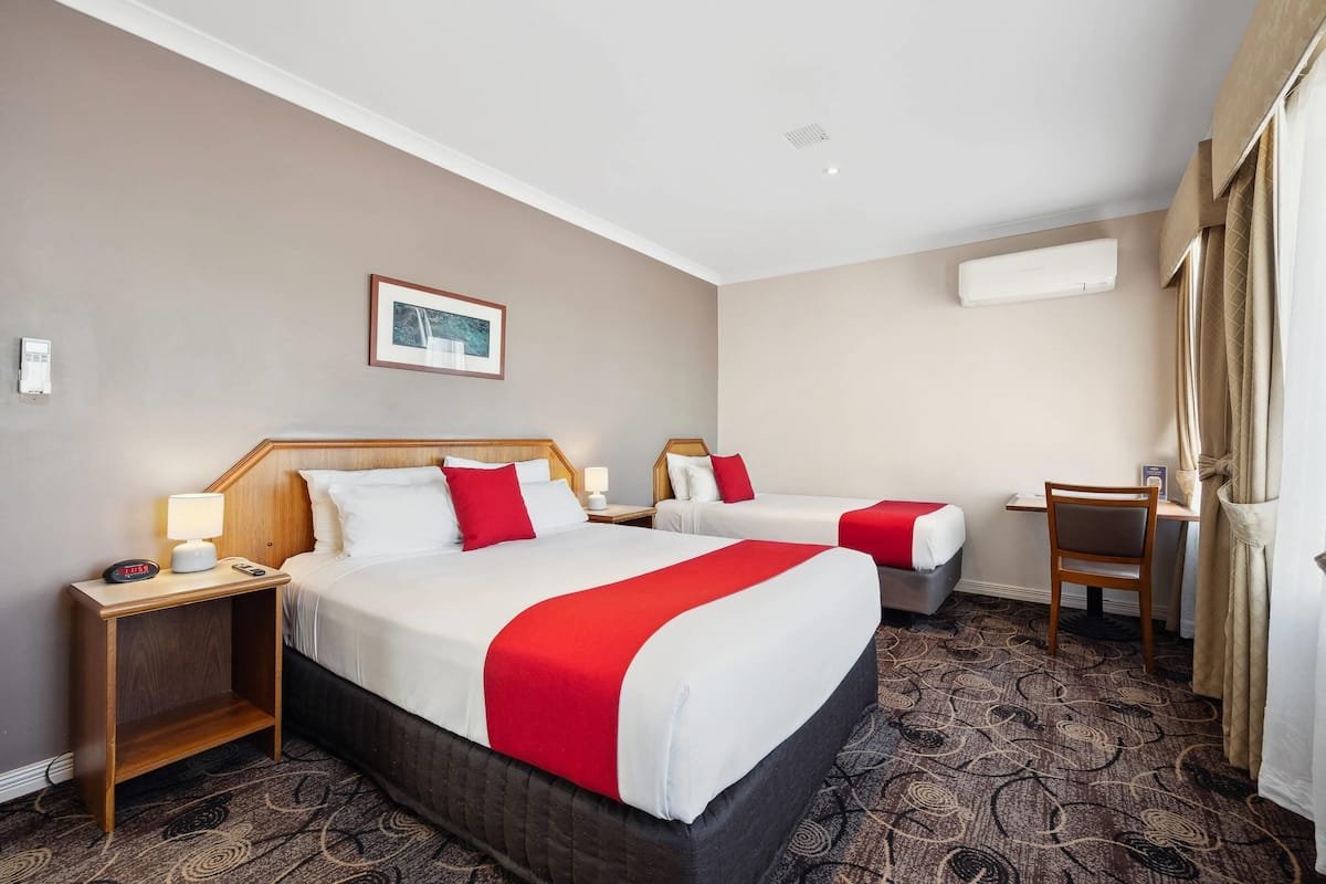 A hotel room with two beds, a desk, a chair, a nightstand with a lamp, and an air conditioning unit.