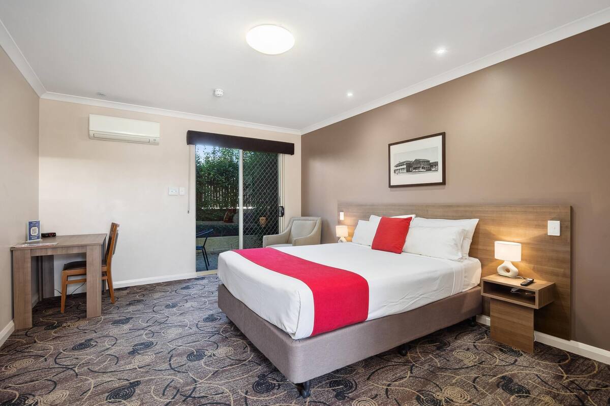 A hotel room with a bed, a desk, a chair, a nightstand, and a window with a view of greenery.