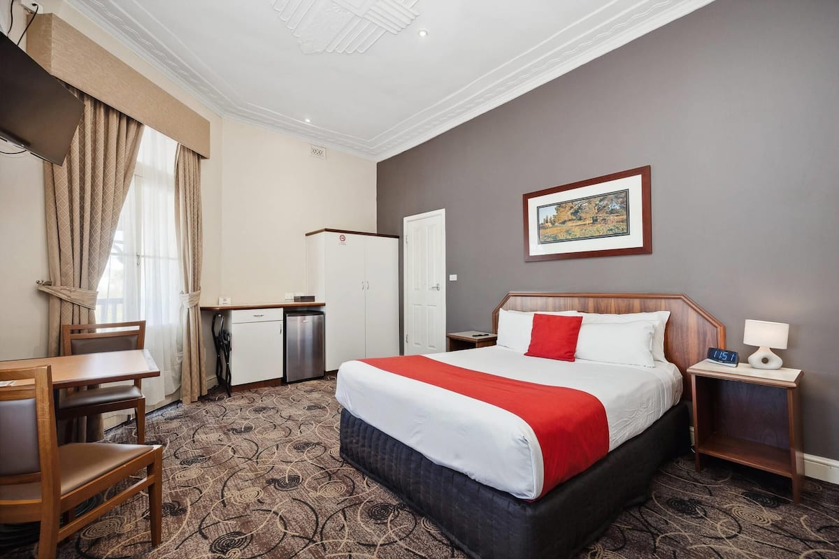 A hotel room with a bed, a desk, a chair, a wardrobe, and a nightstand with a lamp.