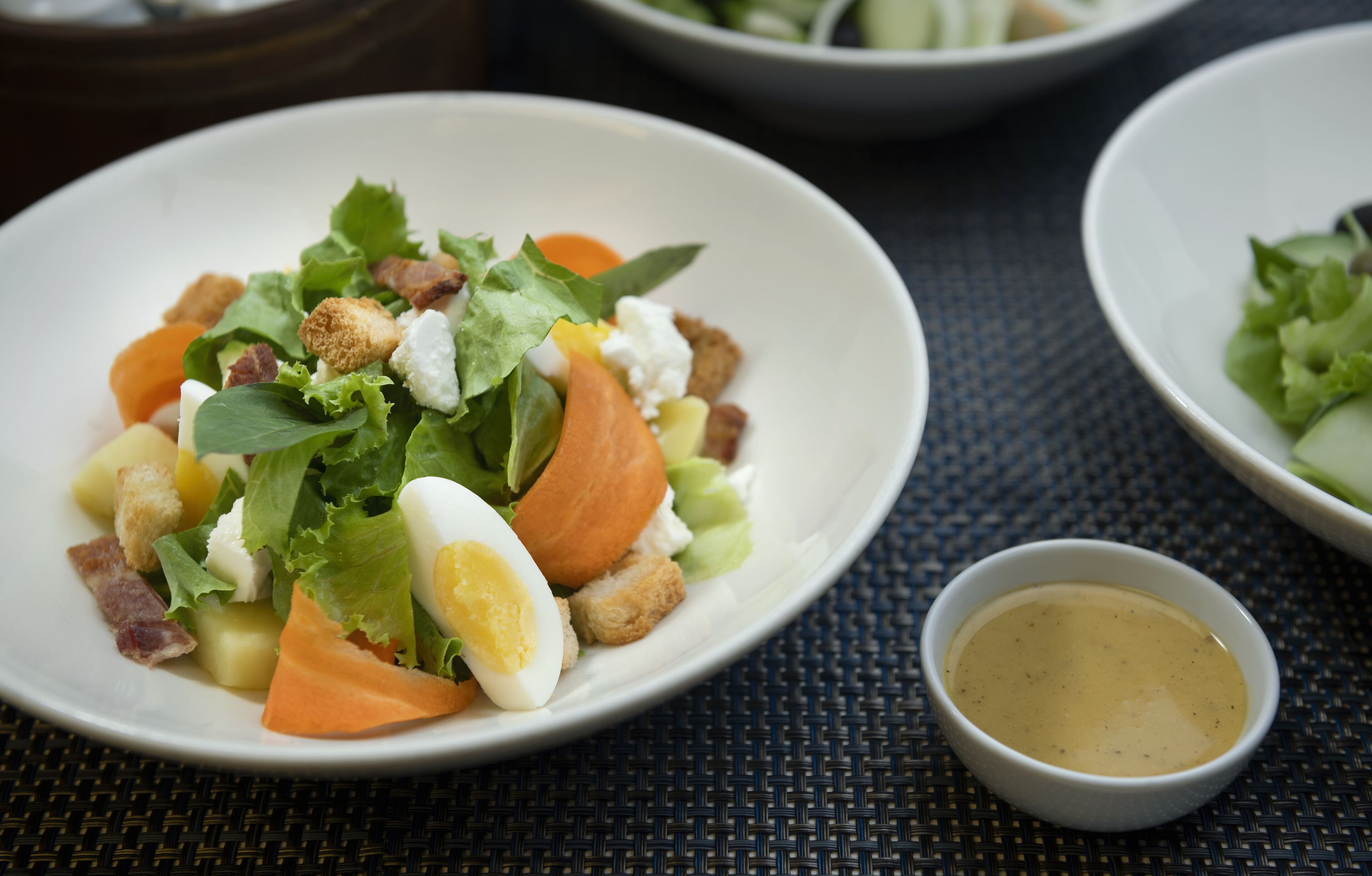 A salad with various ingredients on a white plate, accompanied by a small bowl of dressing.
