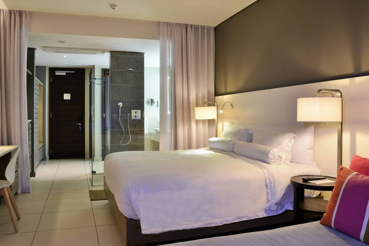A hotel room with a bed, bedside table, lamp, chair, and a desk with a computer.
