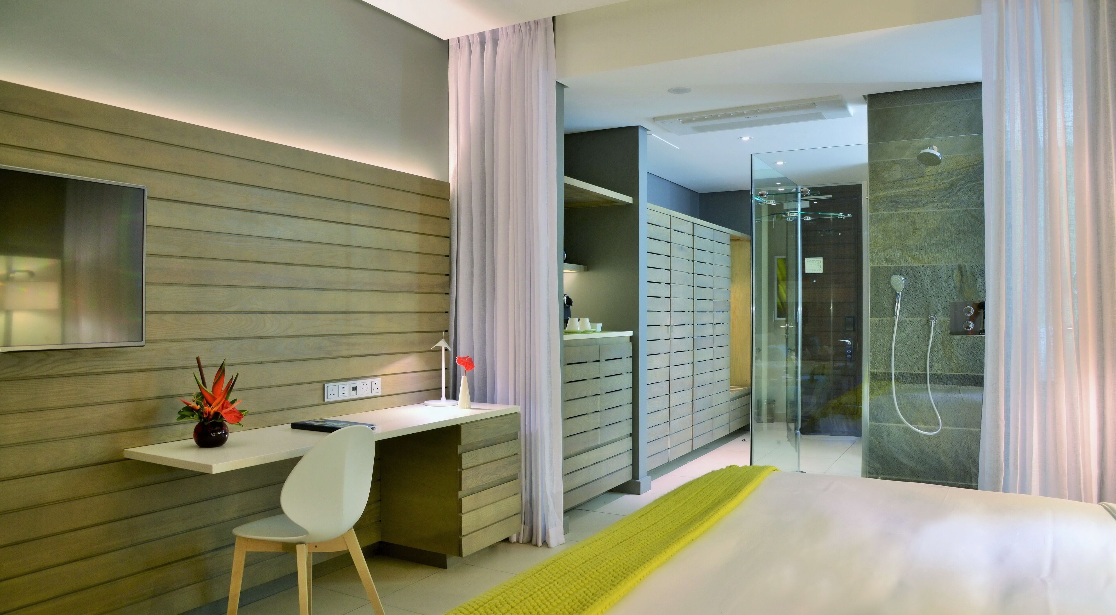 A modern hotel room with a bed, a desk, a chair, a TV, a shower, and a glass-enclosed walk-in closet.