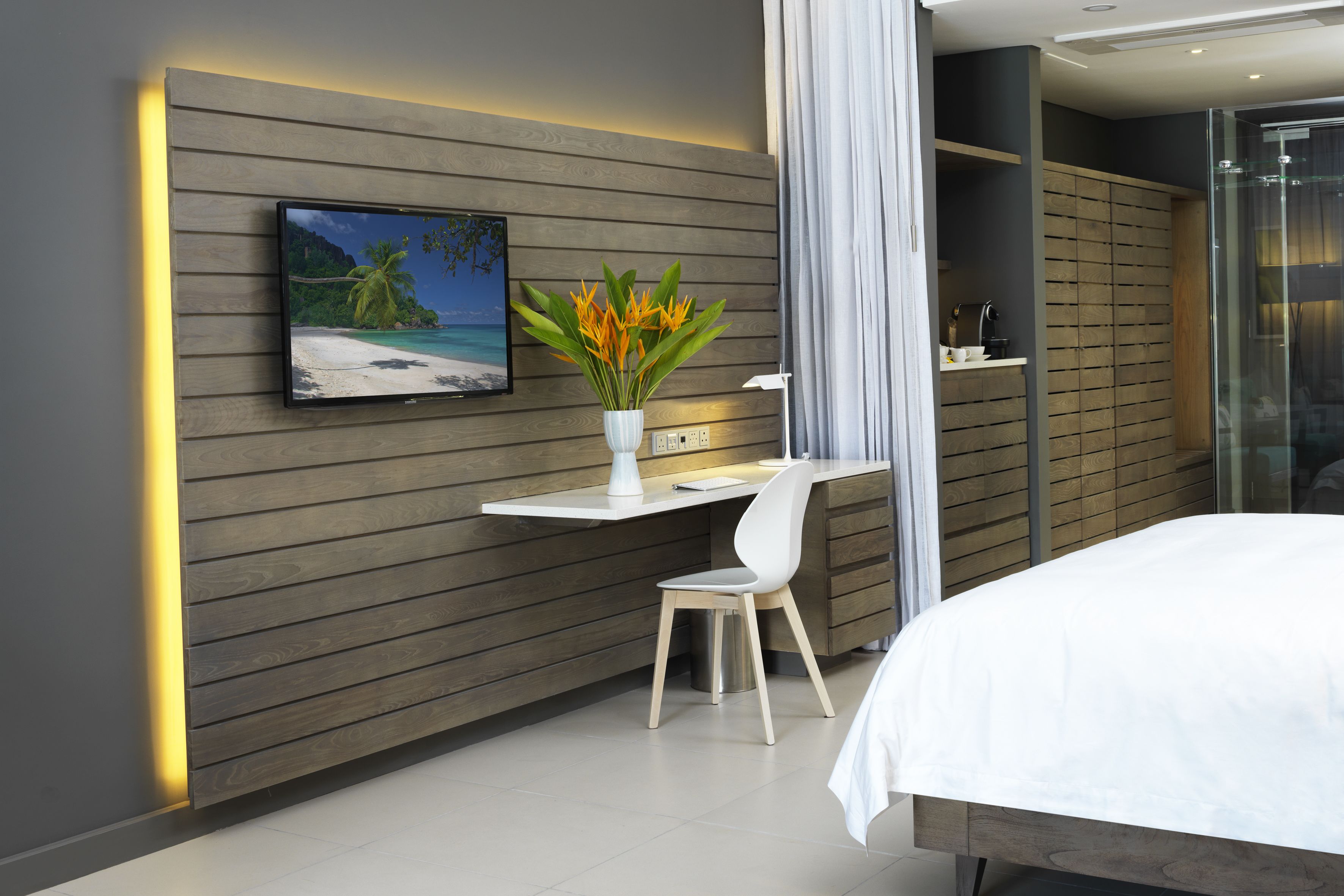A modern hotel room with a wooden panel wall, a desk with a chair, a bed, and a large window with curtains.