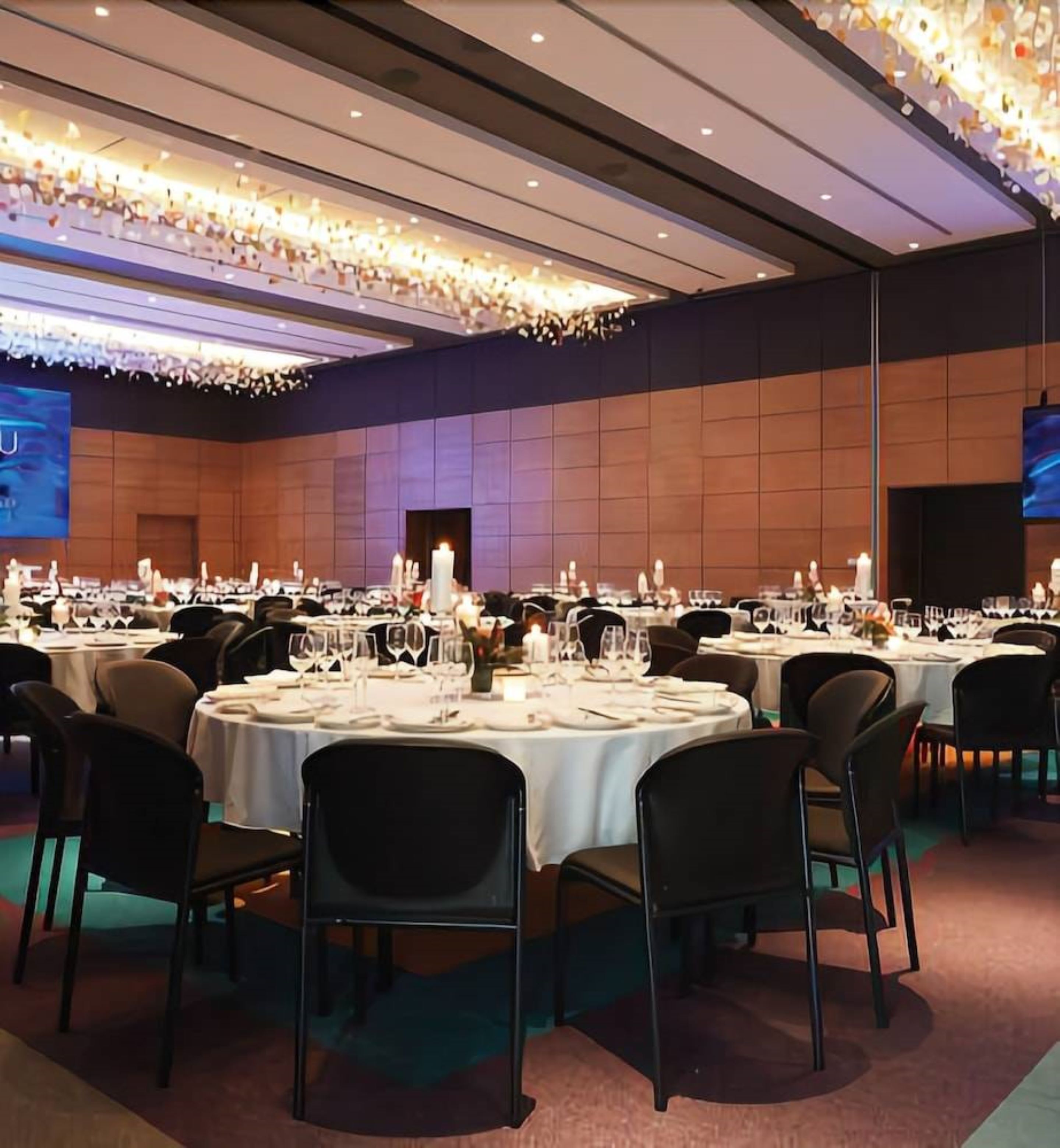 Un salón de banquetes con mesas redondas dispuestas para cenar, una pantalla grande y una araña.