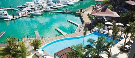A marina with yachts docked, a swimming pool with lounge chairs, and a resort building with a red roof.