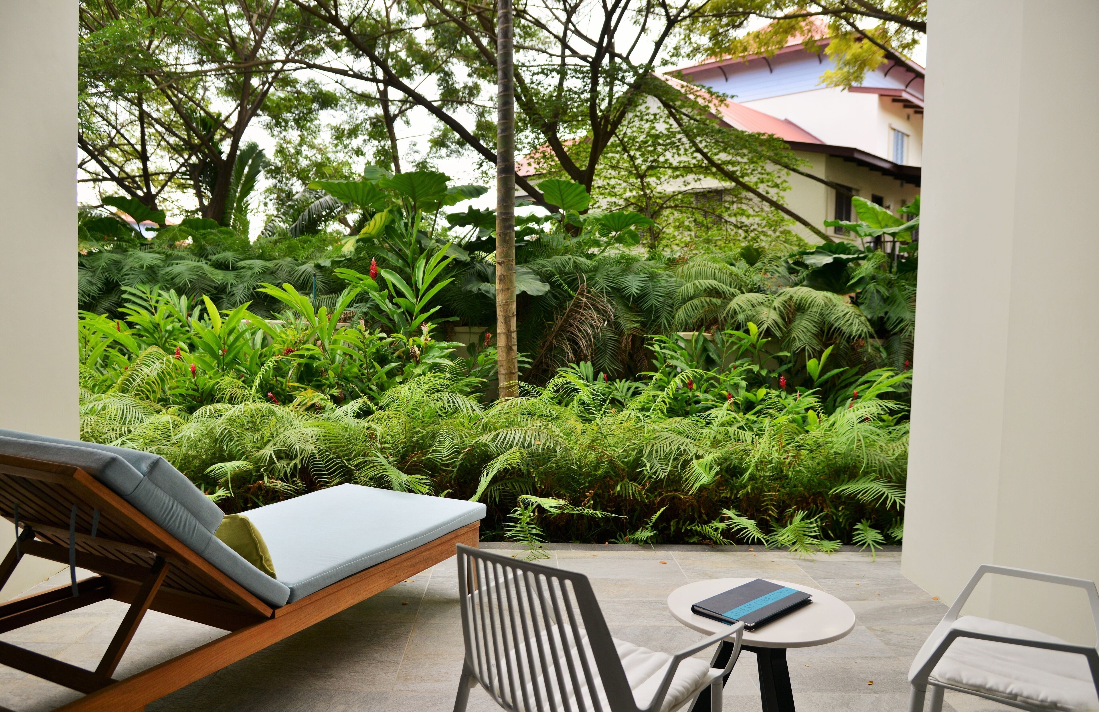 A patio with a lounge chair, a small table, and a chair. The patio is surrounded by lush greenery and trees.