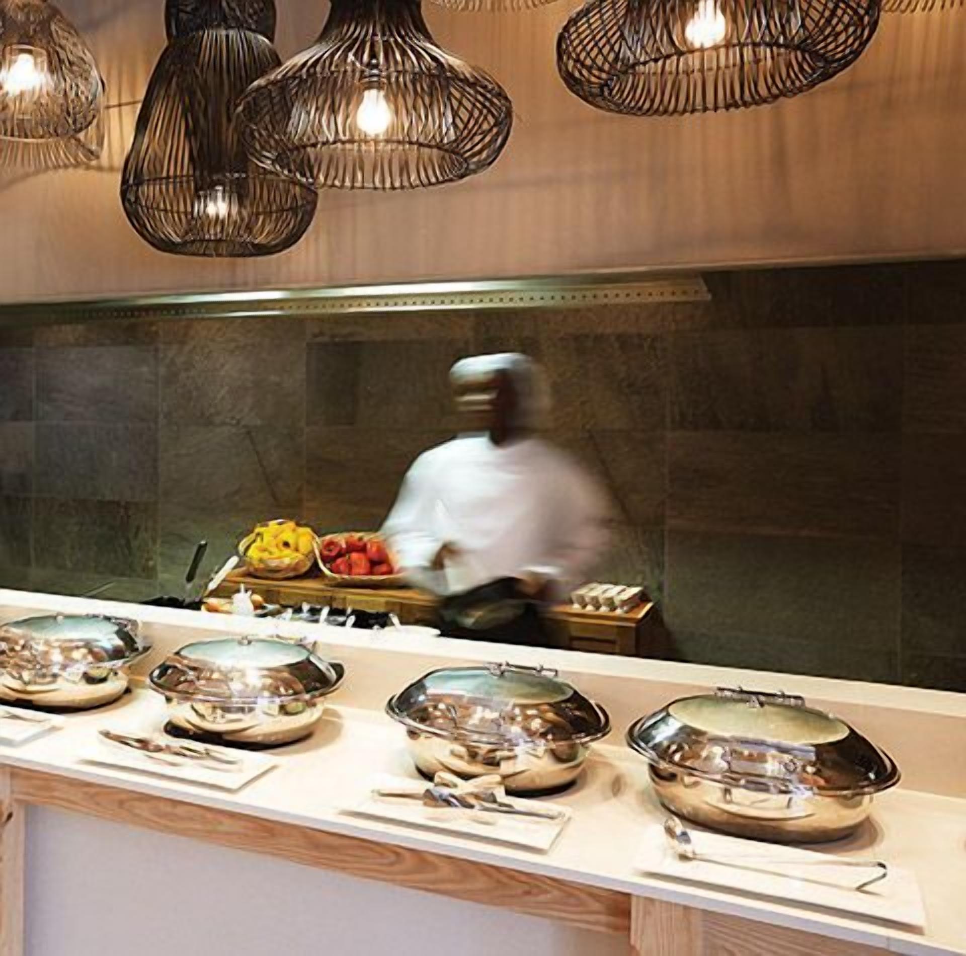 Una cocina con un chef preparando comida, una barra de buffet con platos cubiertos y lámparas colgantes.