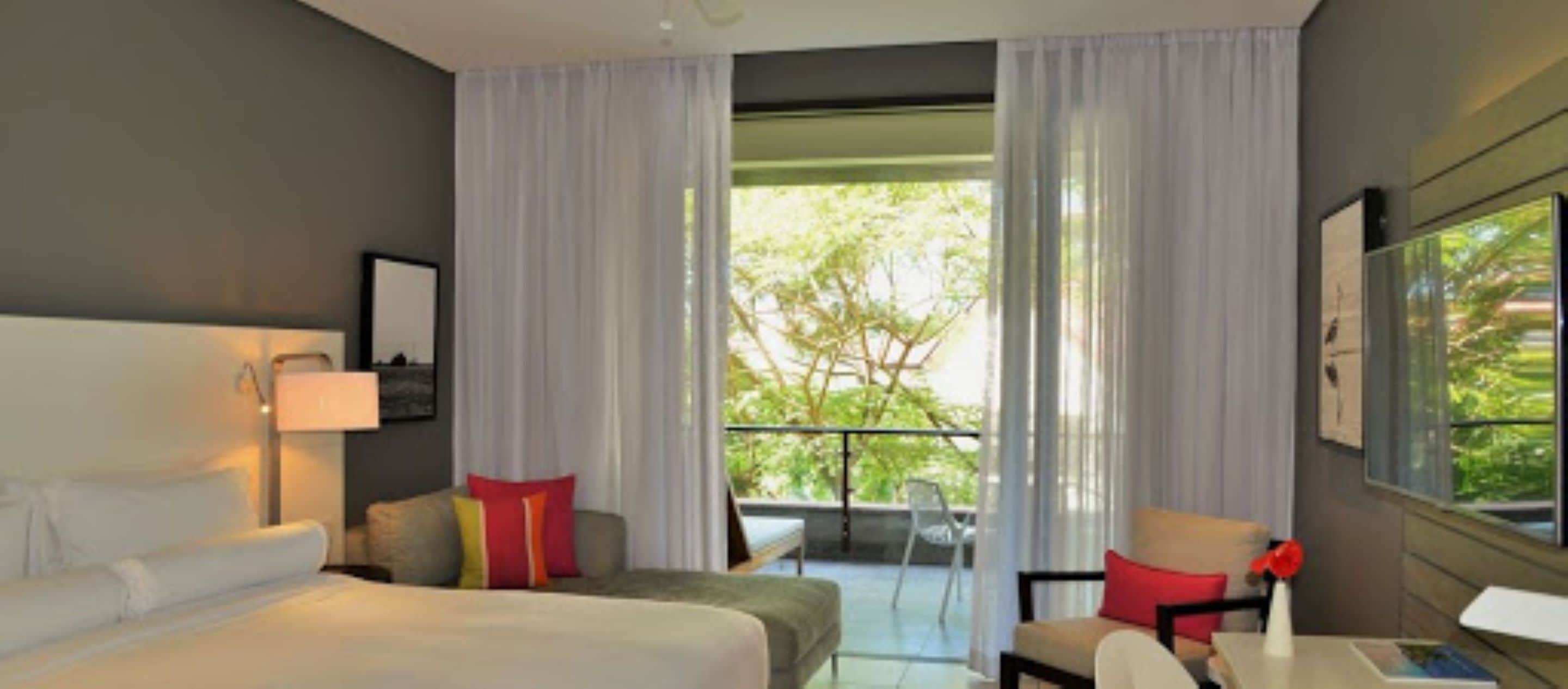 Une chambre d’hôtel avec un lit, un bureau, une chaise et un balcon donnant sur la verdure.