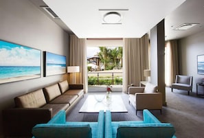 Una habitación de hotel moderna con un amplio ventanal que ofrece vistas al océano, una zona de estar con sofá y sillones, y un pequeño balcón con mobiliario exterior.