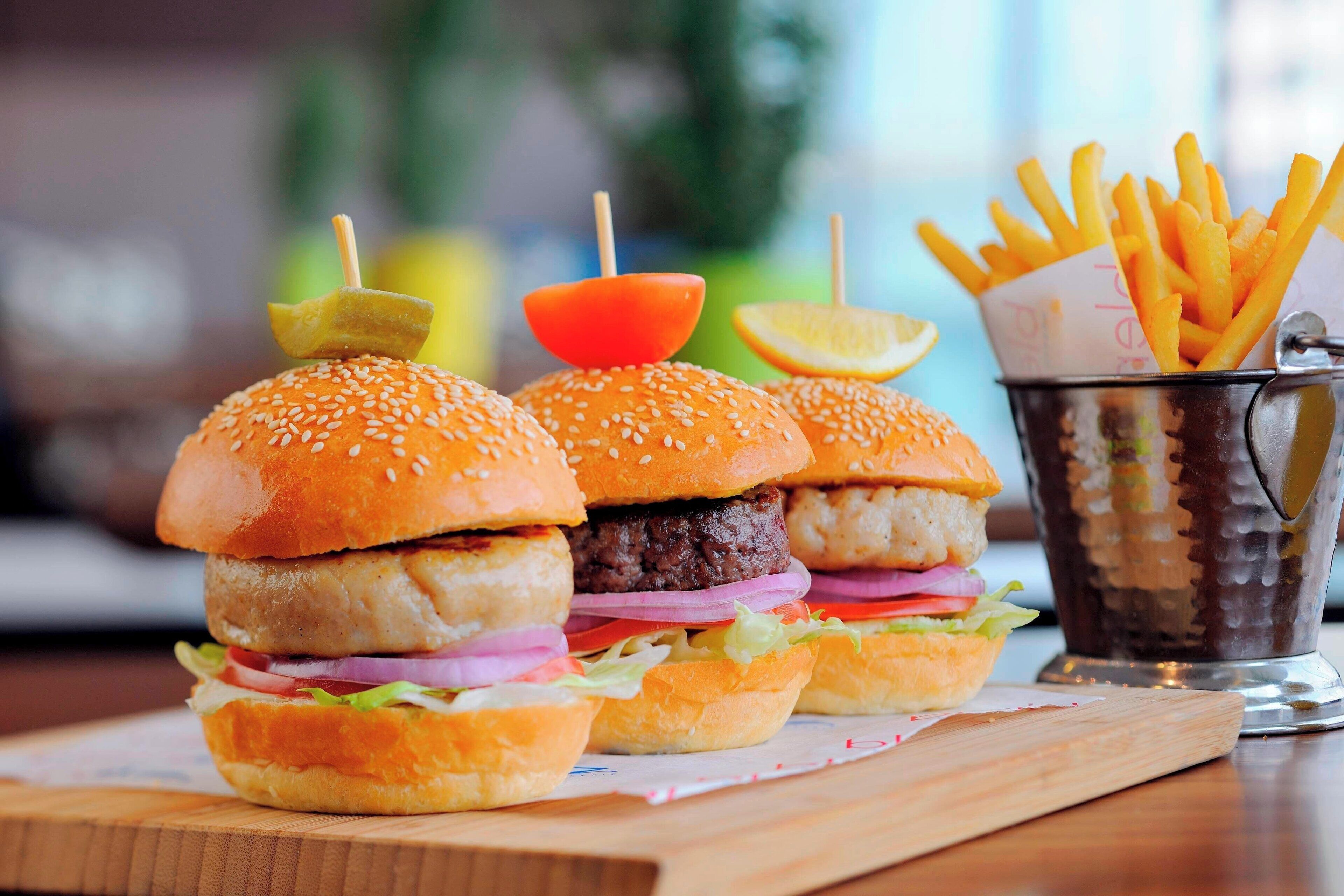 Three burgers with sesame seed buns and toothpicks, a basket of fries, and a drink in a metal cup on a wooden board.