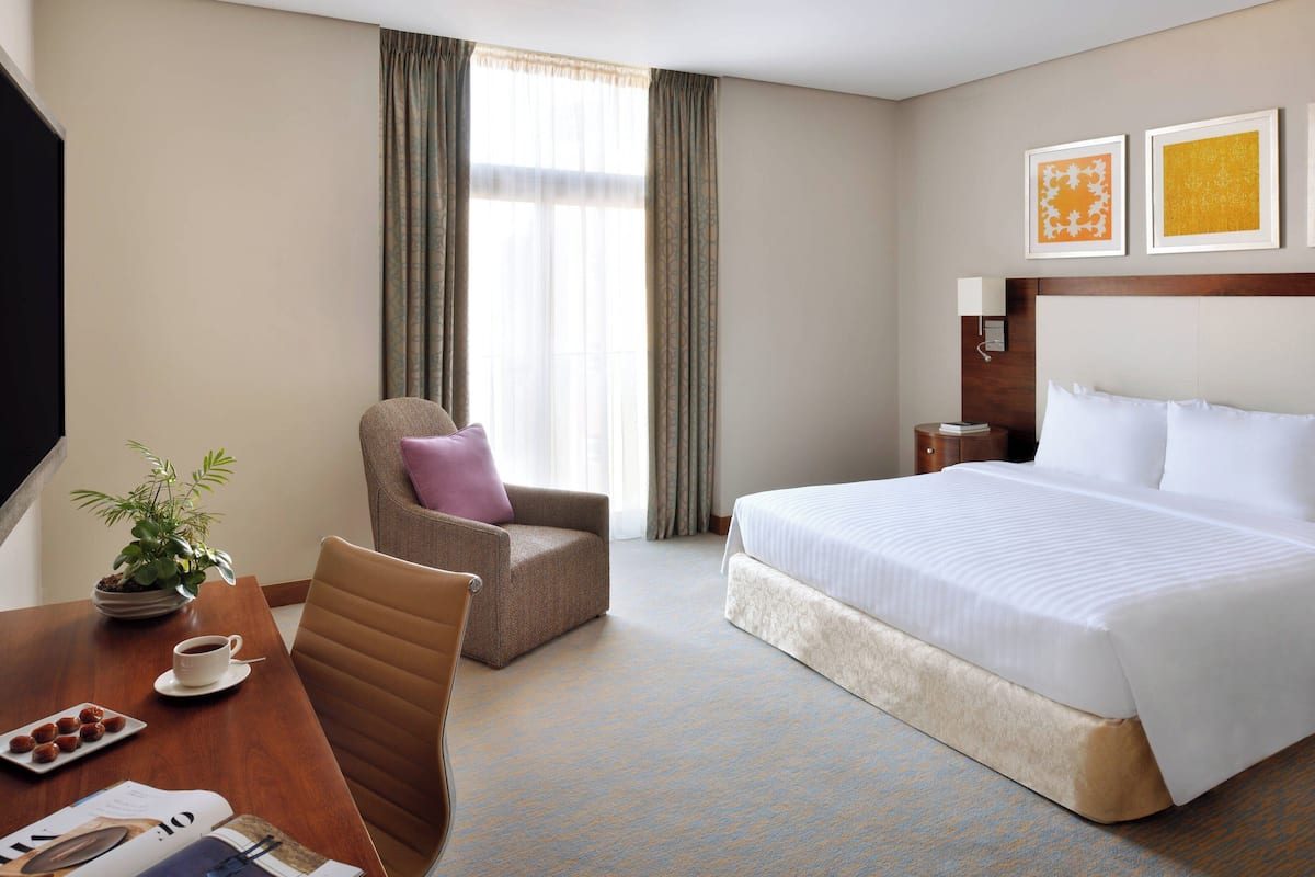 A hotel room with a bed, a chair, a table with a plant and a cup, and a window with curtains.