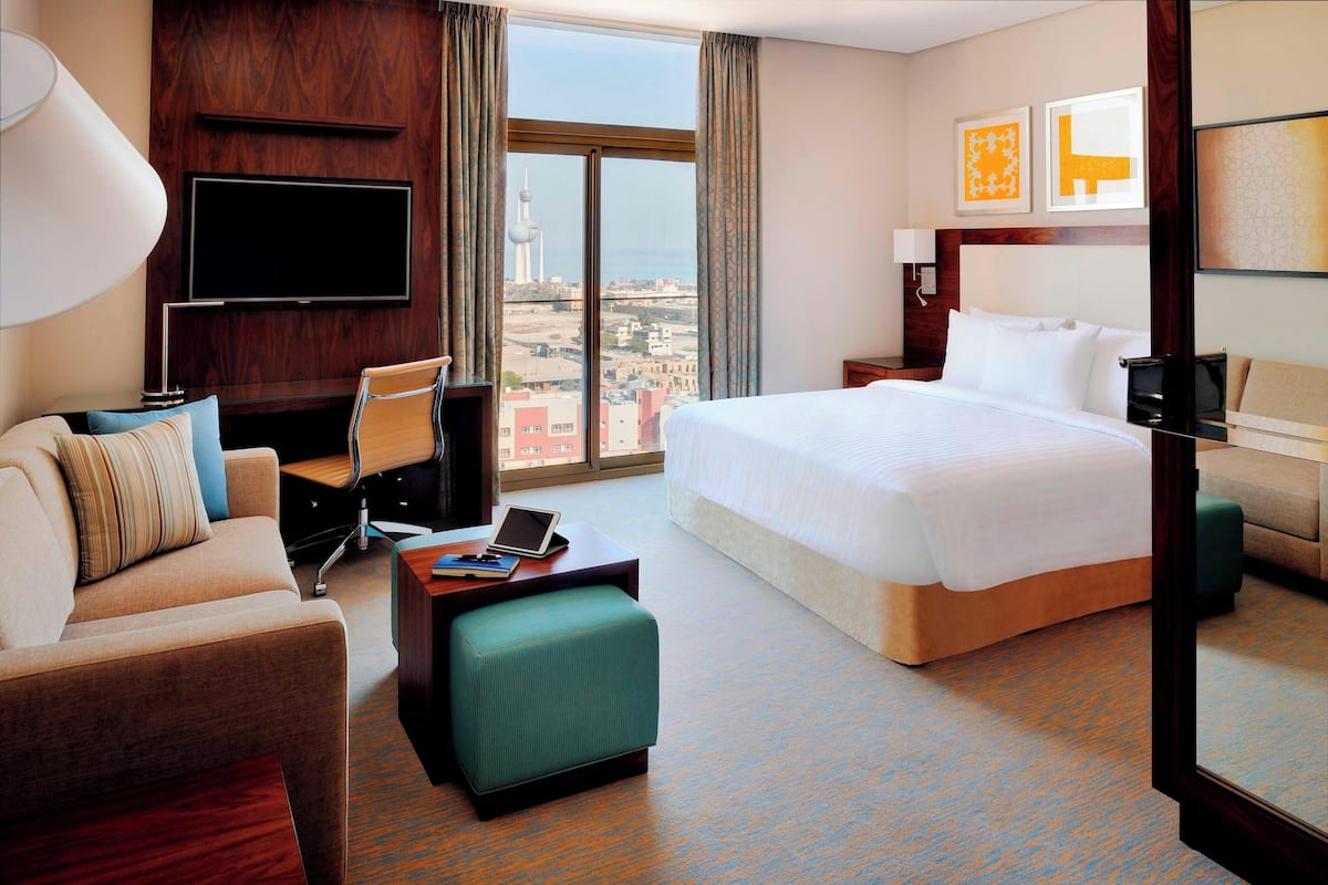 A hotel room with a large bed, a sofa, a desk with a lamp, a TV, and a view of a cityscape.