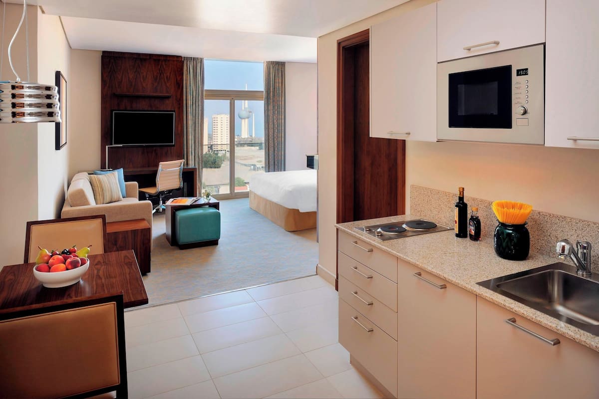 A hotel room with a kitchenette, a dining area, a sofa, a TV, and a view of the city.