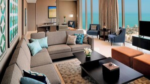 Una sala de estar moderna con un sofá seccional, una mesa de centro y un televisor. Hay un comedor con mesa y sillas, y una vista al océano a través de grandes ventanales.