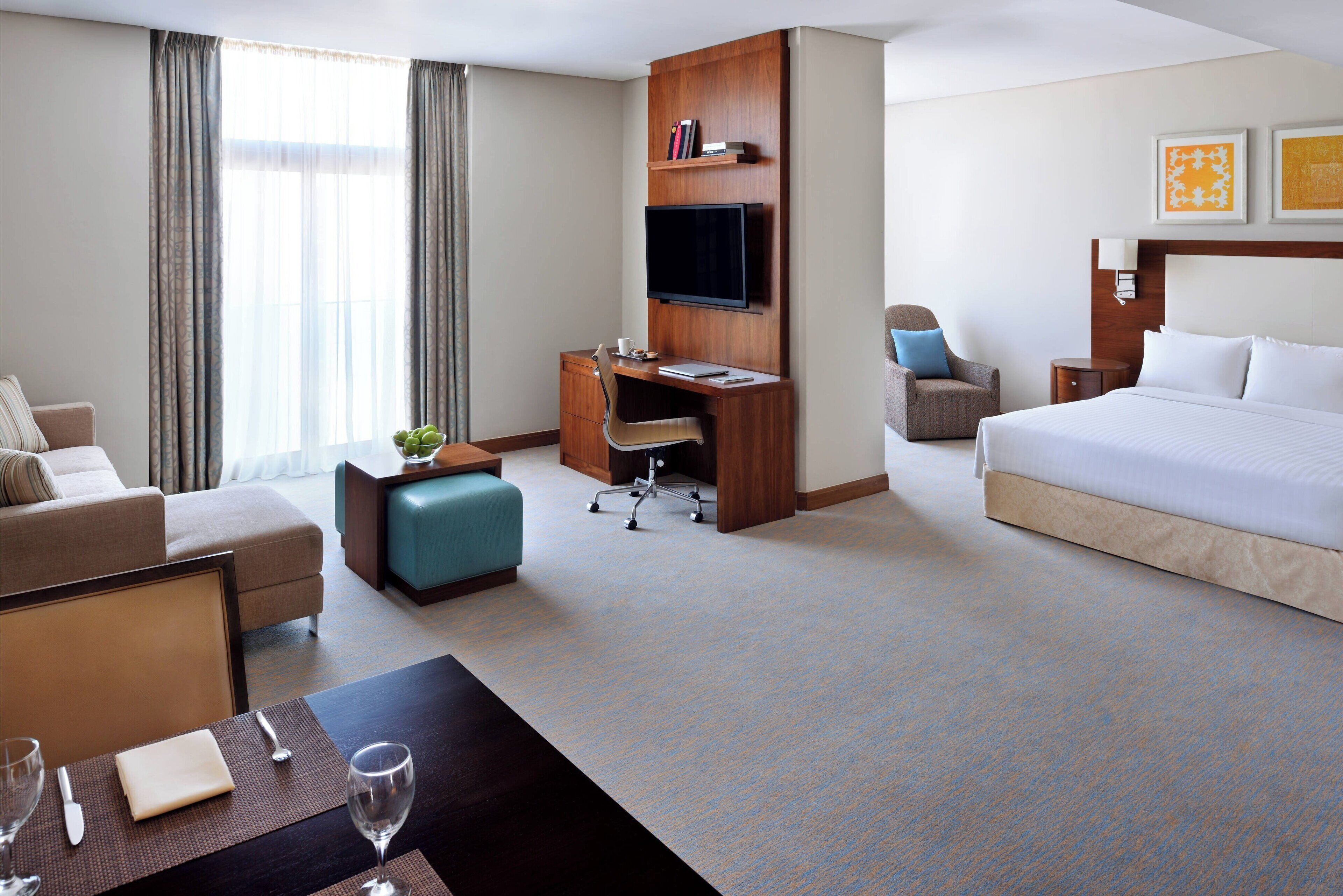 A hotel room with a bed, a desk with a chair, a sofa, a TV mounted on the wall, and a window with curtains.