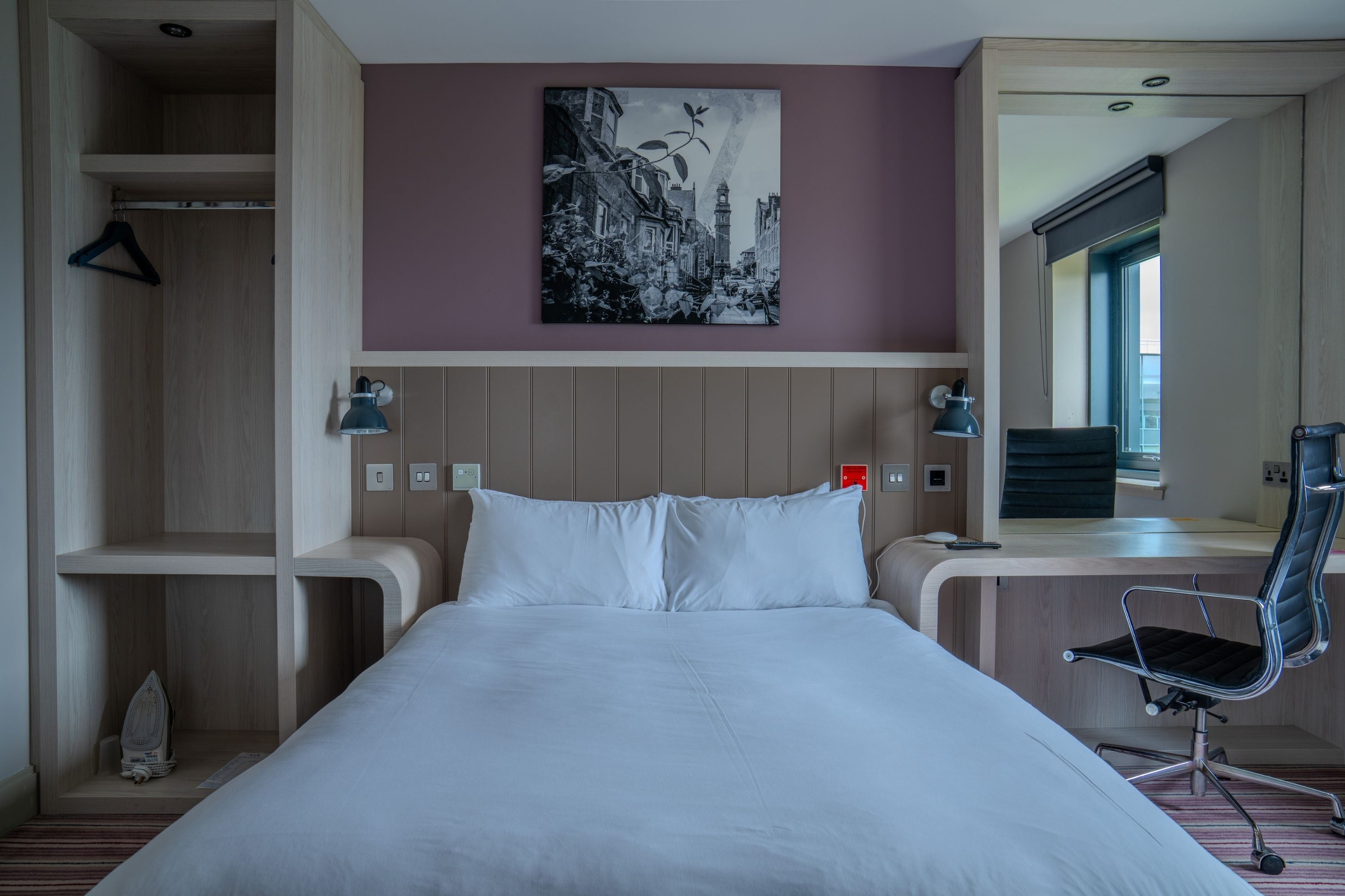 A hotel room with a bed, a desk, a chair, a hanging coat rack, and a painting on the wall.