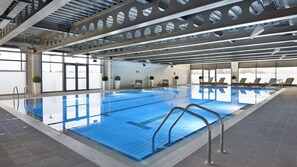 Une grande piscine intérieure à l’eau d’un bleu limpide, entourée de chaises longues et dotée d’un haut plafond orné d’ouvertures circulaires.