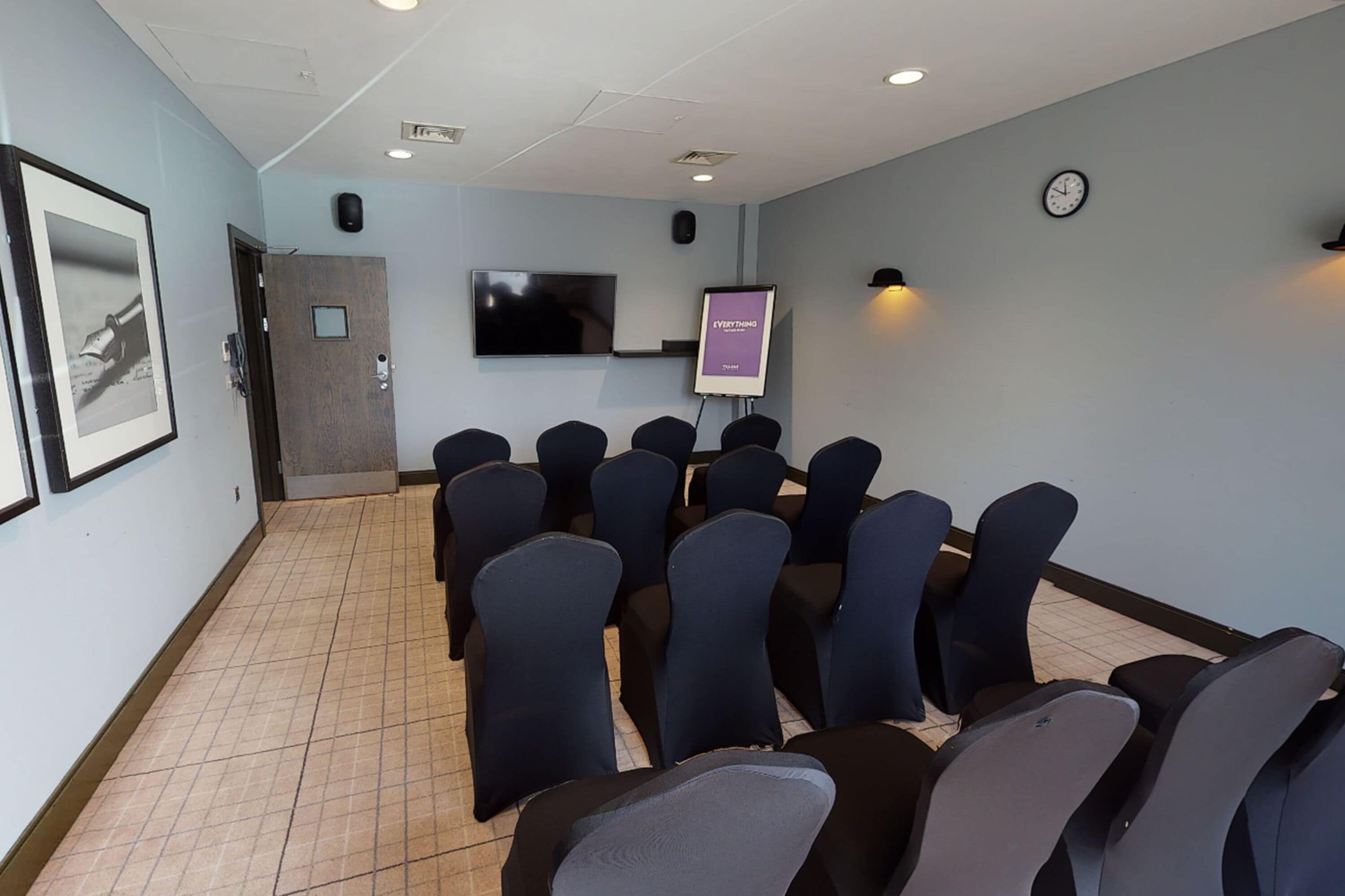 A conference room with rows of chairs, a projector screen, a clock, and a framed picture on the wall.