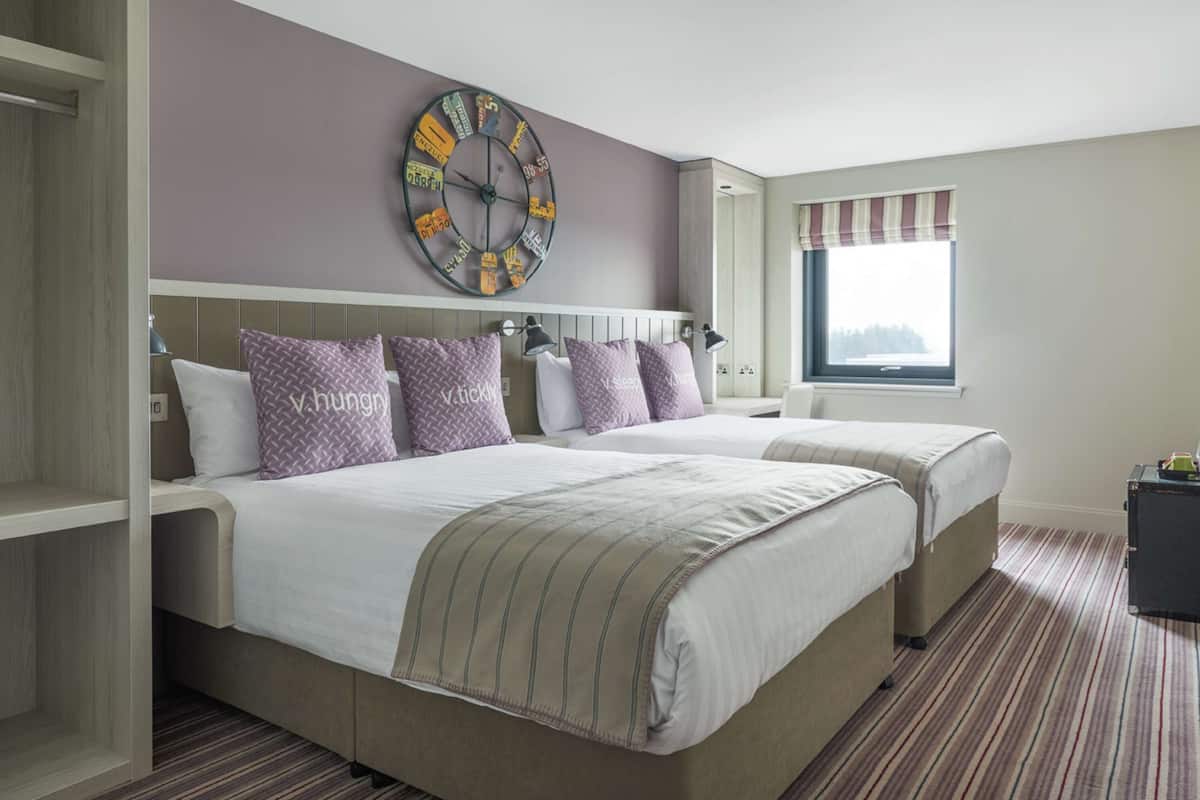 A hotel room with two beds, a large wall clock, and a window with a view.