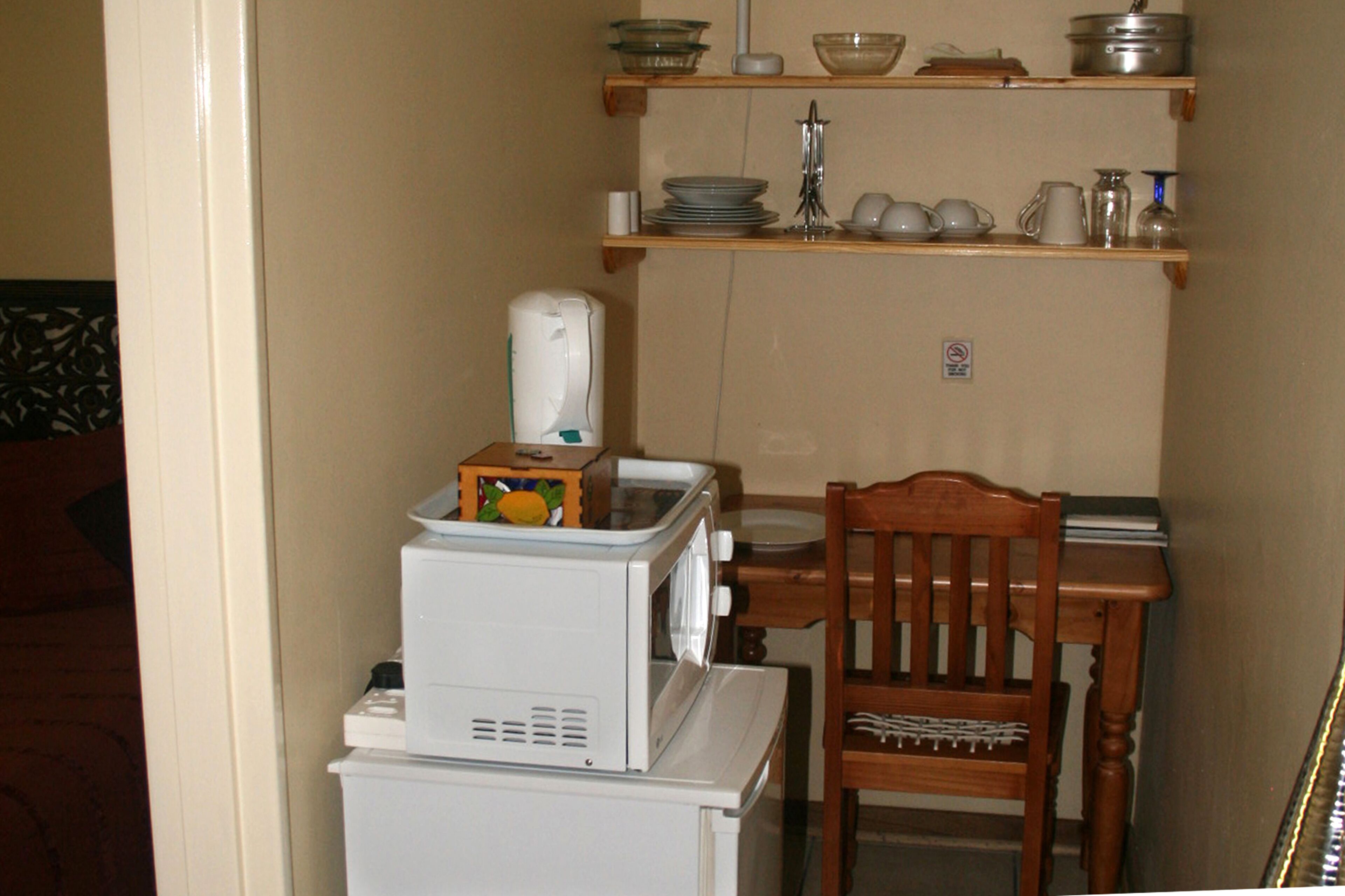 A compact kitchen with a microwave, toaster, and a small refrigerator. There is a wooden chair and a table with shelves above holding dishes and a kettle.