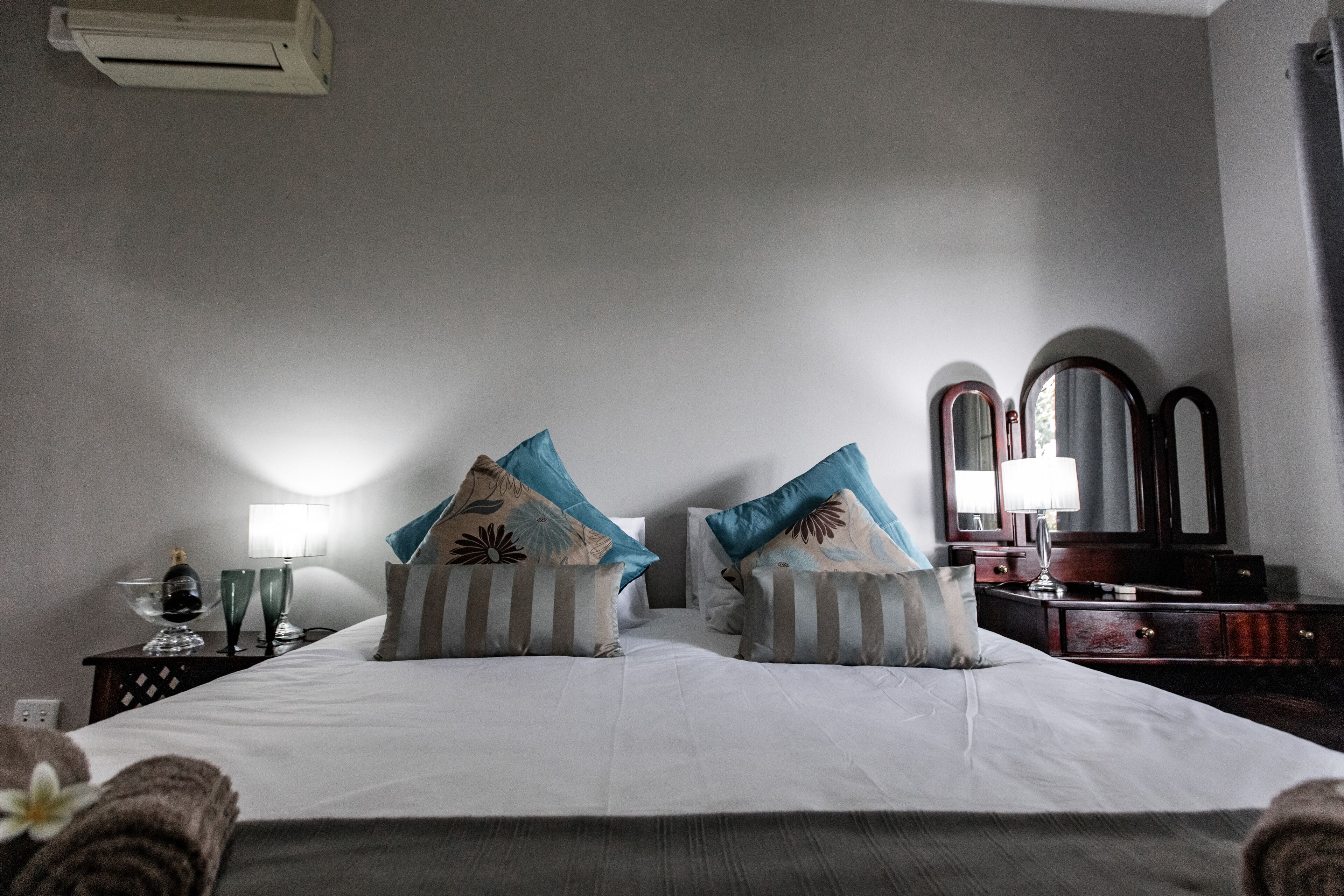 Una habitación de hotel con una cama grande, dos almohadas, una mesita de noche con lámpara, un espejo y un aparador.