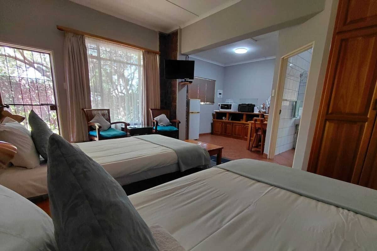 A hotel room with two beds, a TV, a kitchenette, and a view of trees through the window.