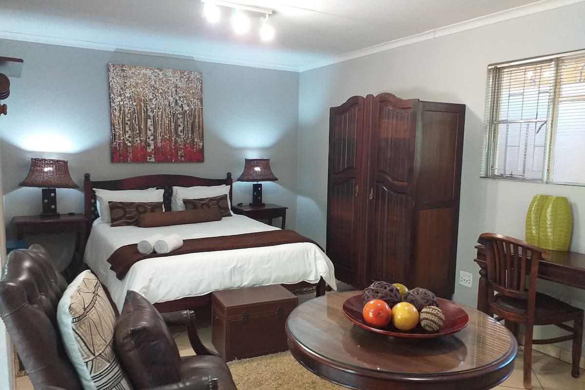 A neatly arranged bedroom with a bed, bedside lamps, a wooden wardrobe, a dining table with fruit, and a leather armchair.
