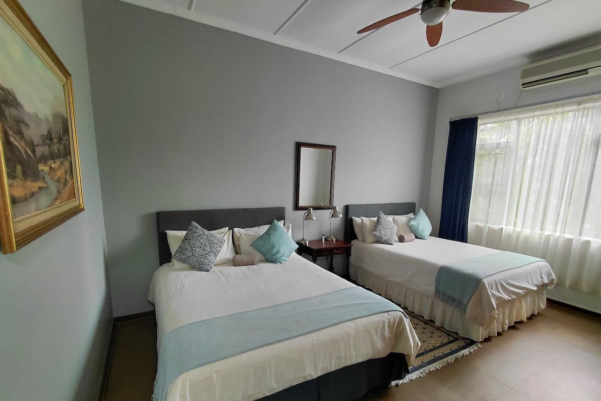 A hotel room with two beds, a ceiling fan, a painting, and a large window with curtains.