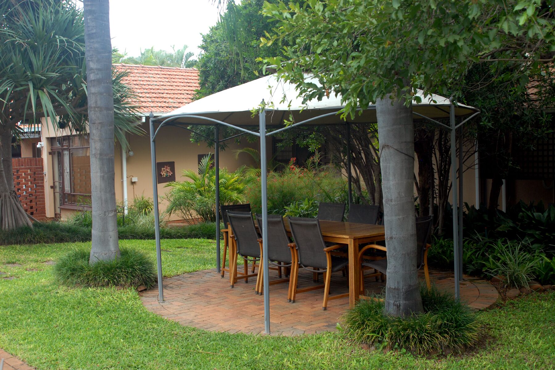 A covered outdoor dining area with a brick patio, surrounded by lush greenery and a building with a red-tiled roof.