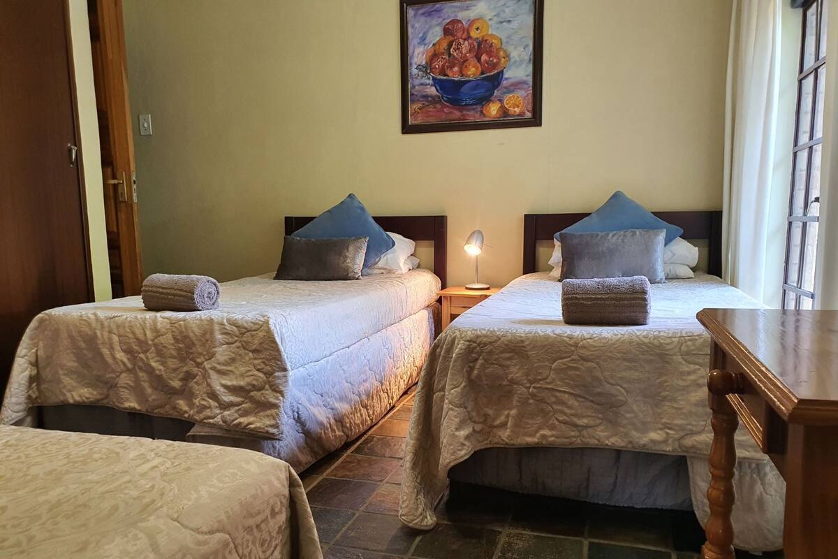 A hotel room with two beds, each with a white quilted bedspread and matching pillows. There is a painting of fruit on the wall above the beds, a nightstand with a lamp between the beds, and a wooden table to the right.