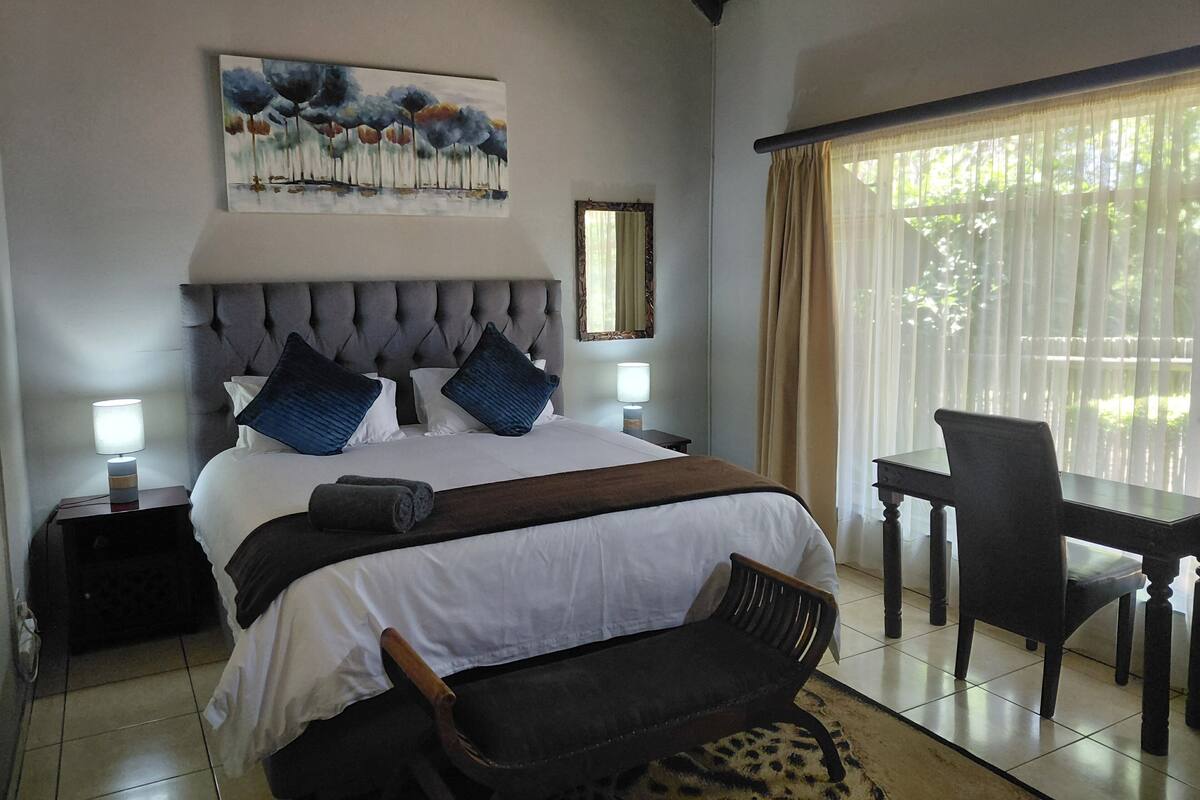 A bedroom with a large bed, a desk, a chair, a painting on the wall, and a window with curtains.