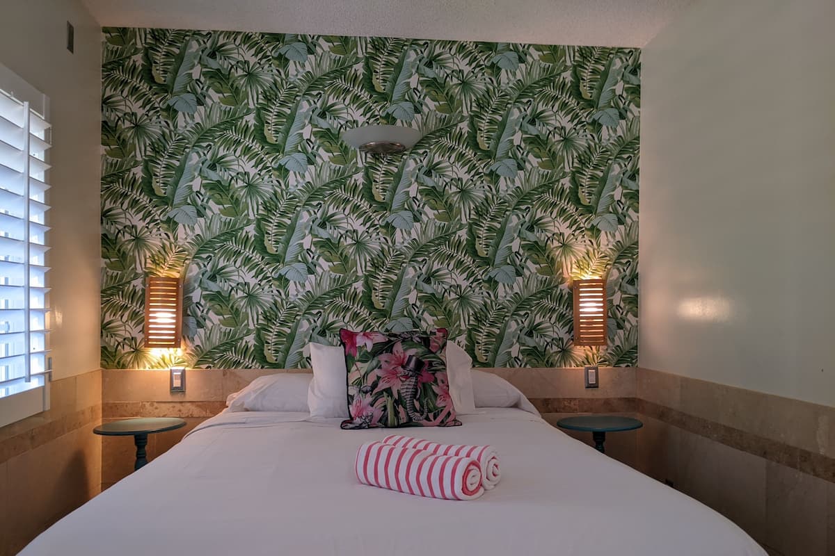 Habitación de hotel con una cama grande, dos mesitas de noche, un cojín decorativo y una pared con un diseño de hojas tropicales.
